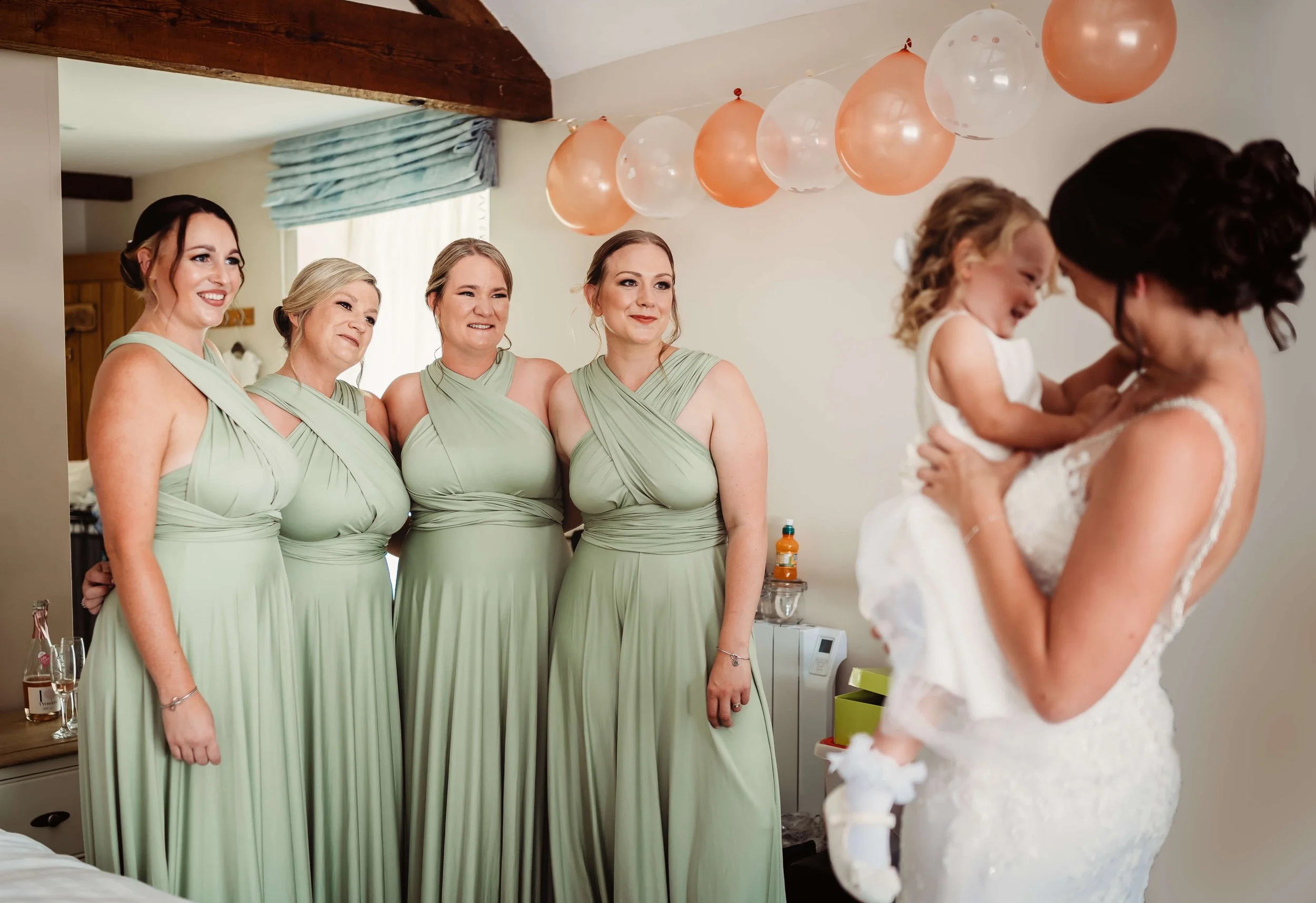 A bride holding a laughing young girl, possibly her daughter, while four women in matching light green bridesmaid dresses look on in a decorated room with balloons.