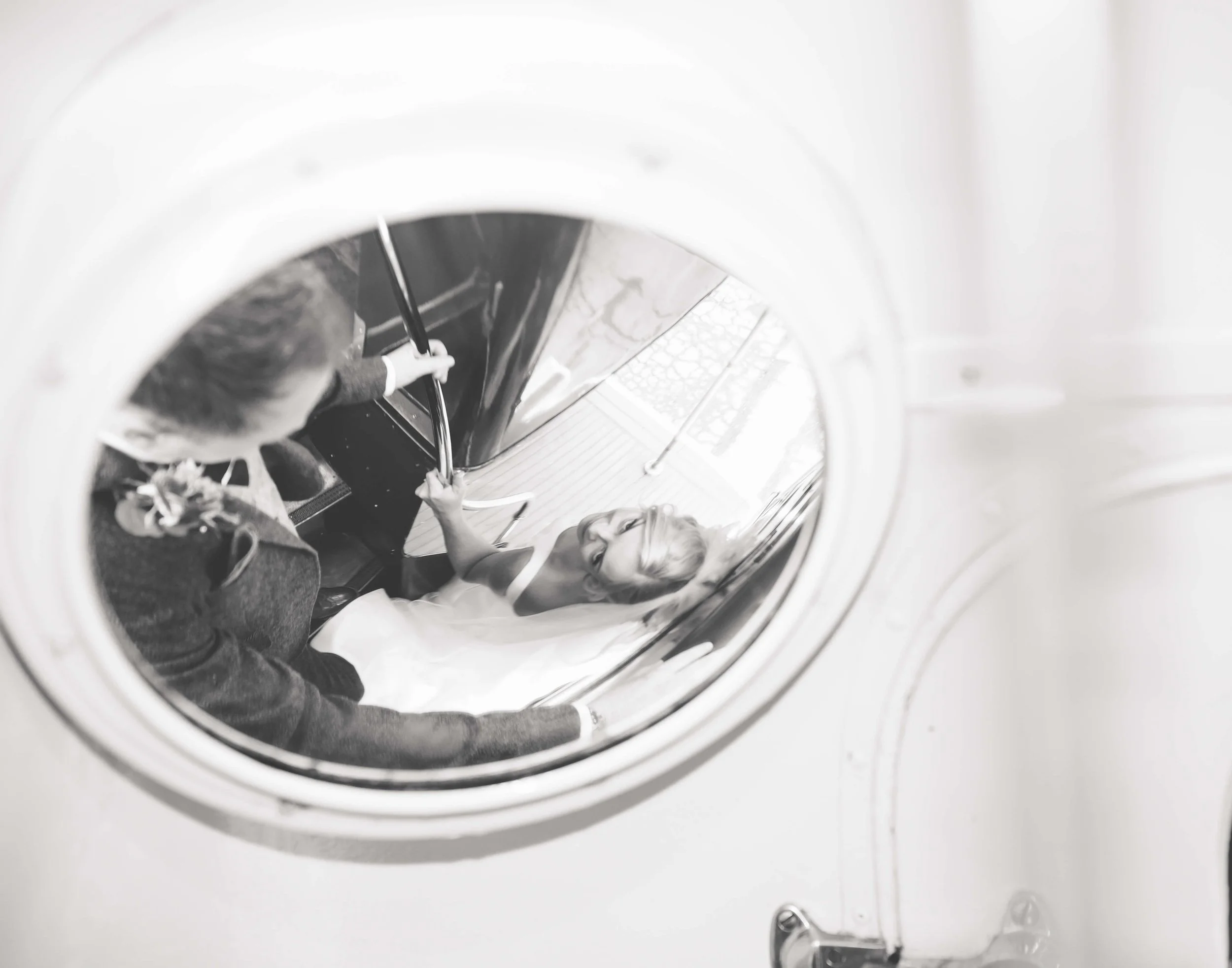 A woman in a wedding dress and veil is seen through the door of a front-loading washing machine, with another person inside, holding her hand.