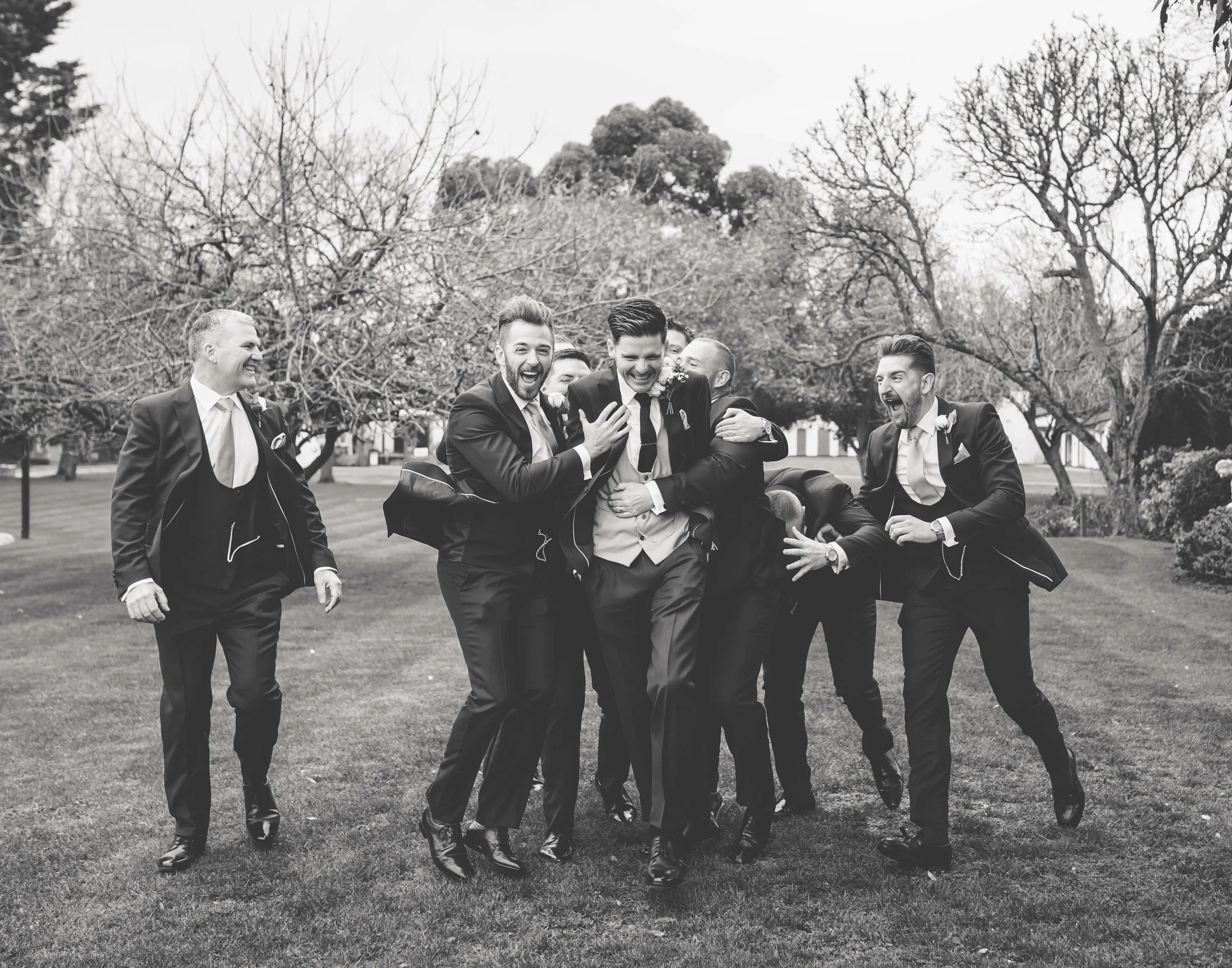 Group of men dressed in suits, celebrating and laughing outdoors on a cloudy day.