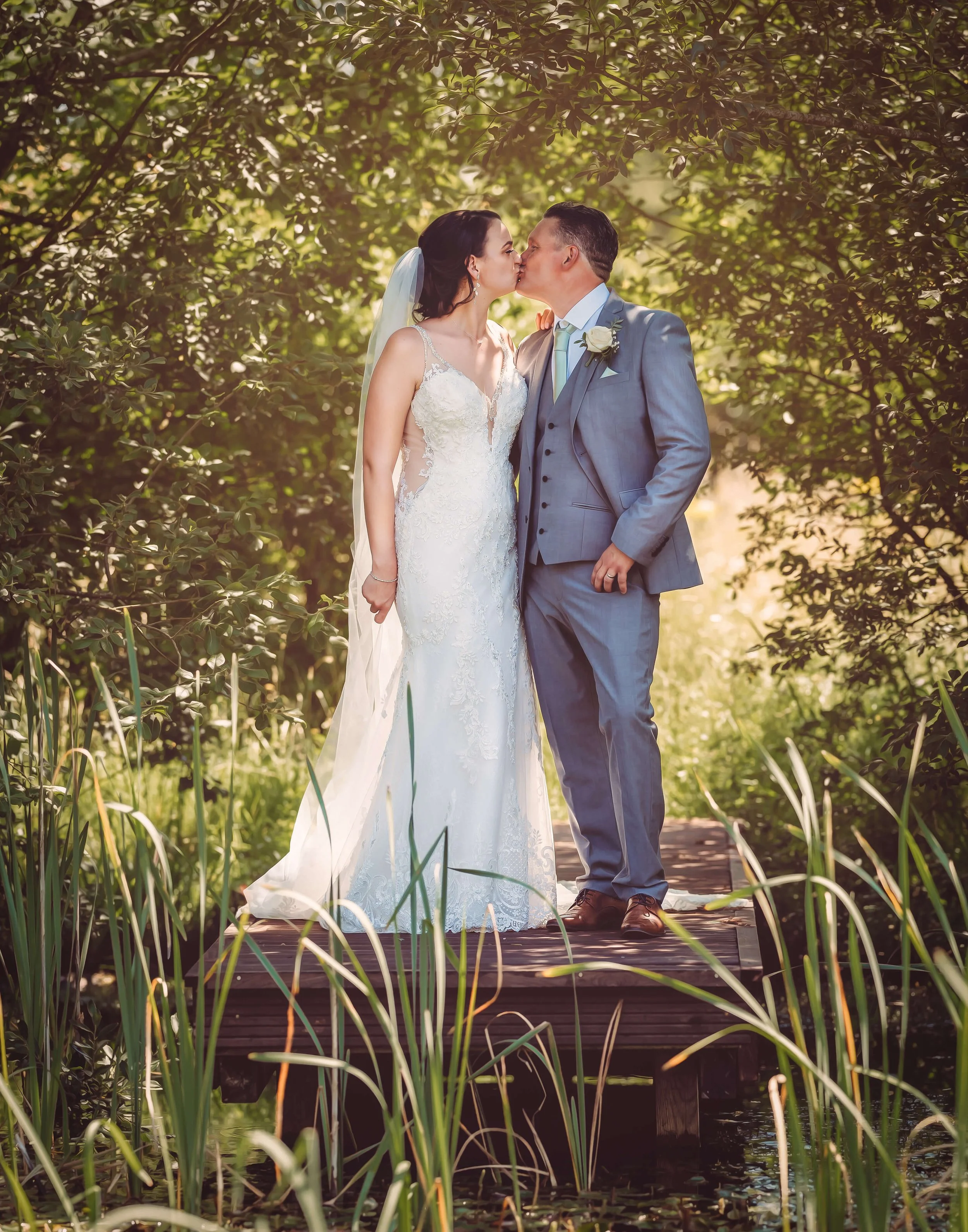 A newlywed couple sharing a kiss on a wooden dock surrounded by greenery and tall grass in a natural outdoor setting.