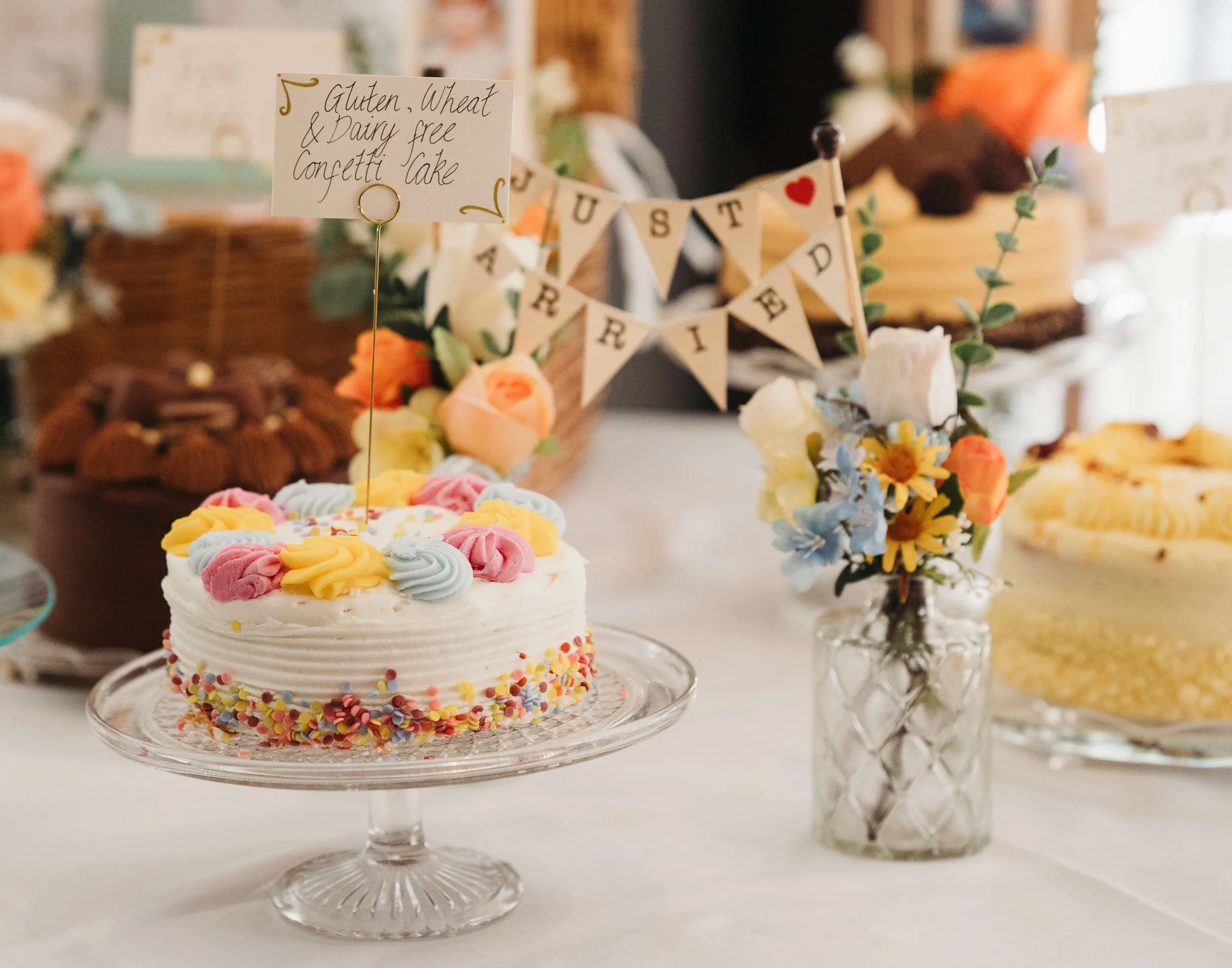 A small birthday cake with pastel colored piped flowers and rainbow sprinkles on a glass cake stand, with a sign on top that reads 'Gluten, Wheat & Dairy free Confetti Cake'; behind, a vase with assorted flowers and a banner with the name 'Aureled' a