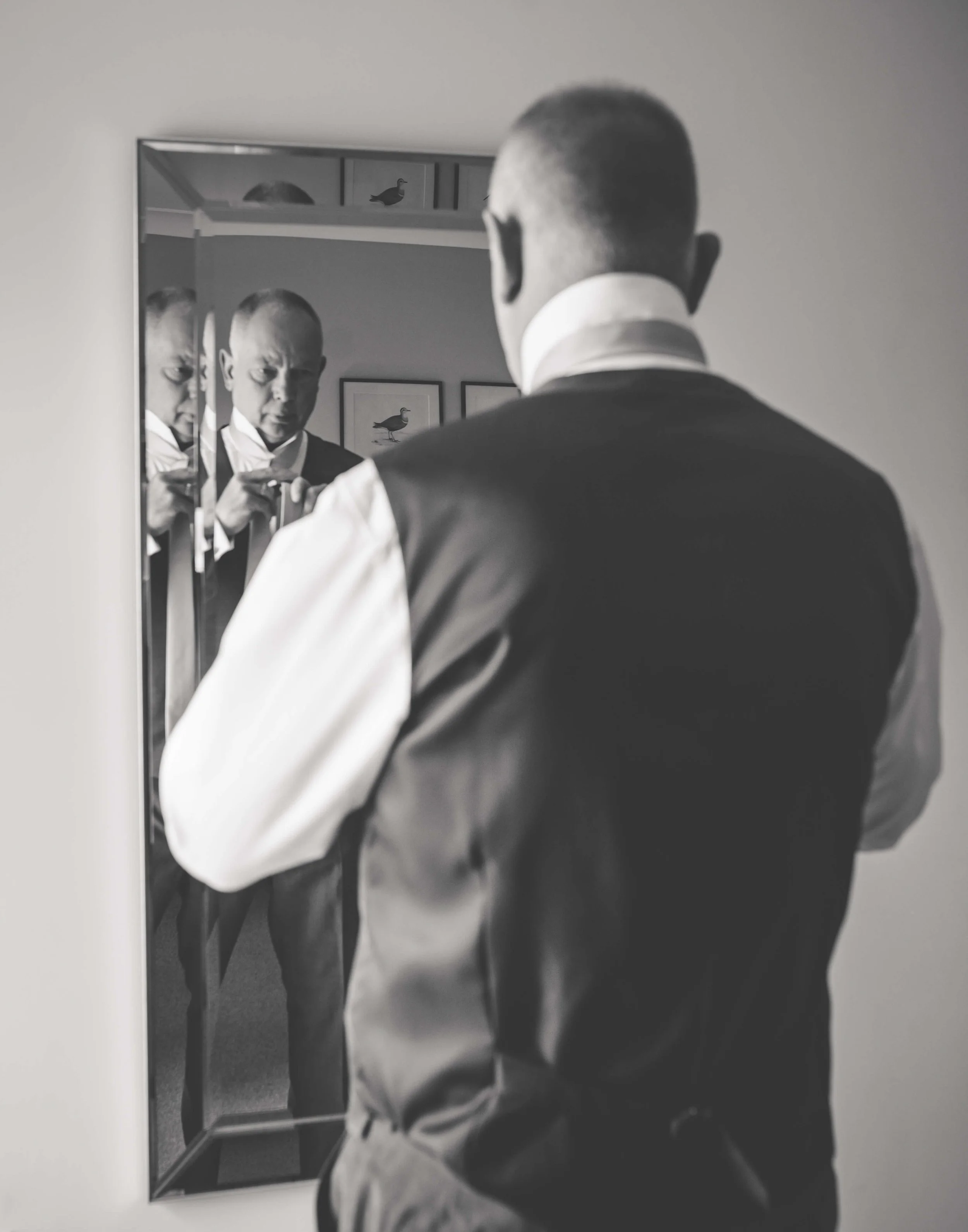 A man in formal attire looking into a mirror, with his reflection adjusting his cufflinks, in black and white.