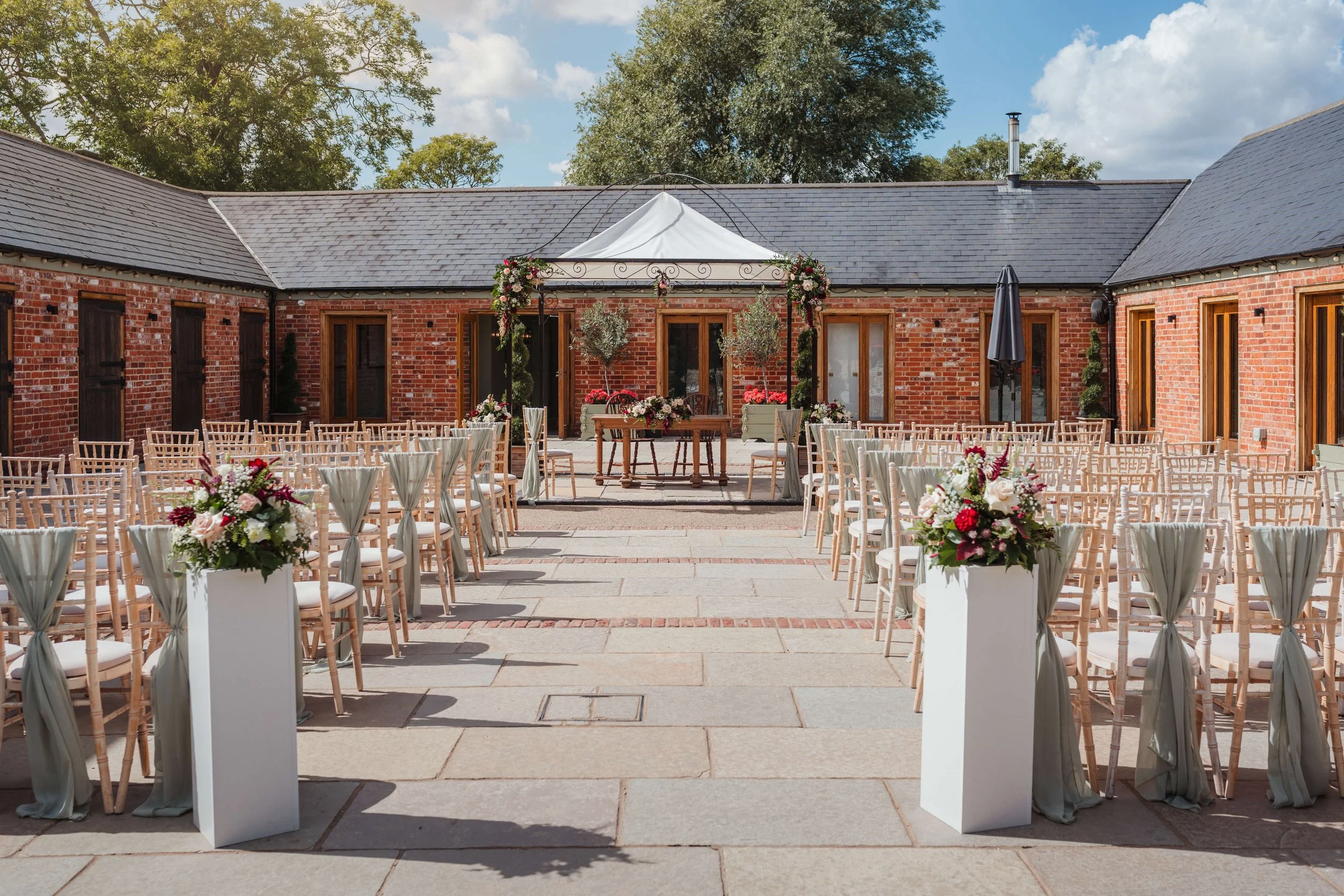 Outdoor wedding ceremony setup on a paved patio with rows of chairs, floral arrangements, a small stage, a white canopy, and a brick building in the background.