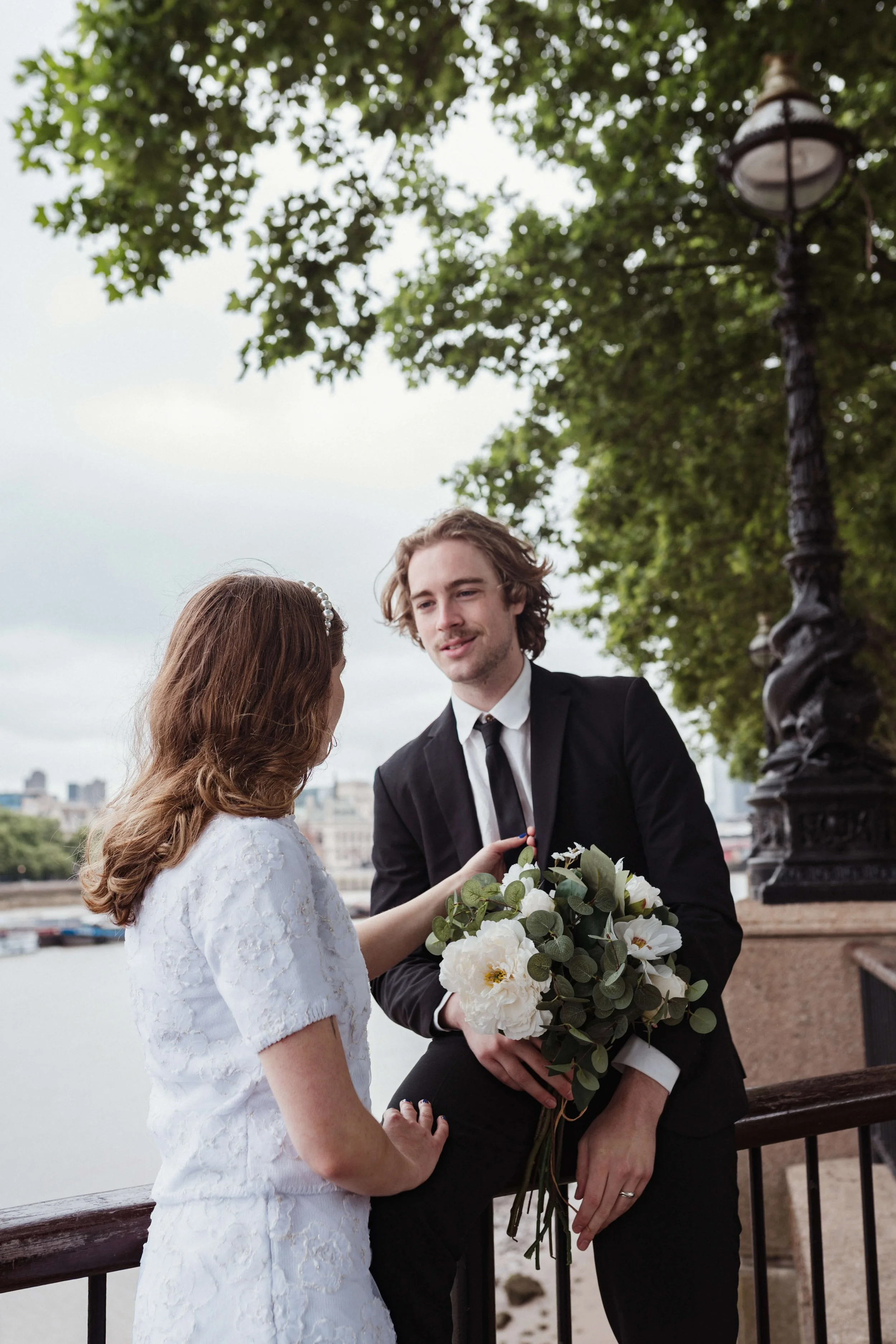 A bride and groom exchanging vows outdoors by a river, with trees and a vintage streetlamp nearby.