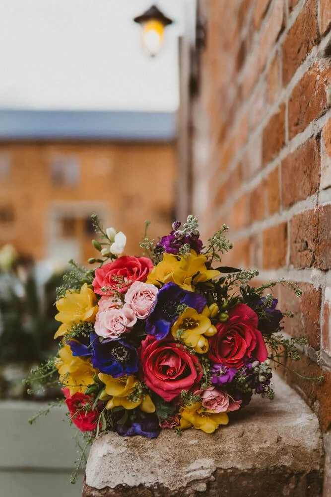 Colorful bouquet of flowers including roses, pansies, and other blossoms, placed on a stone ledge against a brick wall, with a building and a lamp in the background.