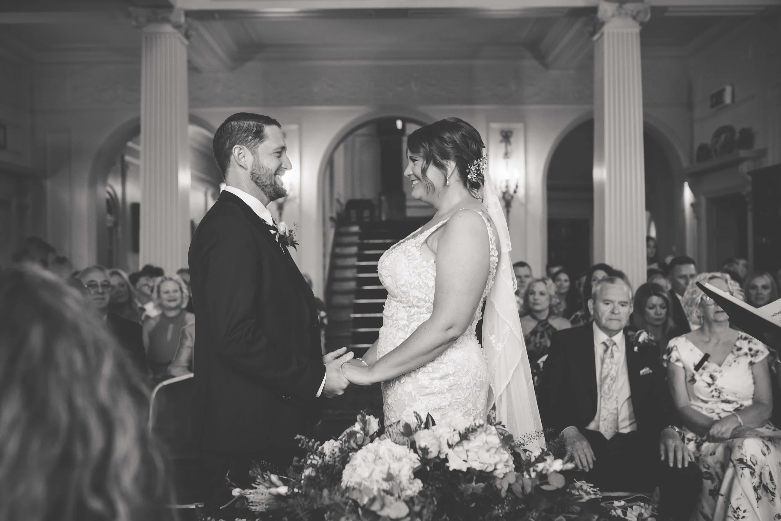A black-and-white photo of a bride and groom standing face to face holding hands at their wedding ceremony inside a grand hall, with guests seated around them.