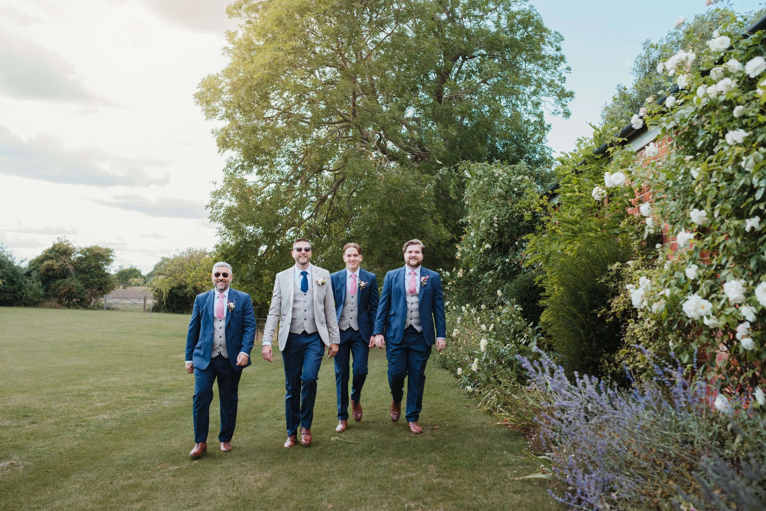 Group of five men dressed in formal suits walking outdoors on green grass, with trees and flowering bushes in the background.