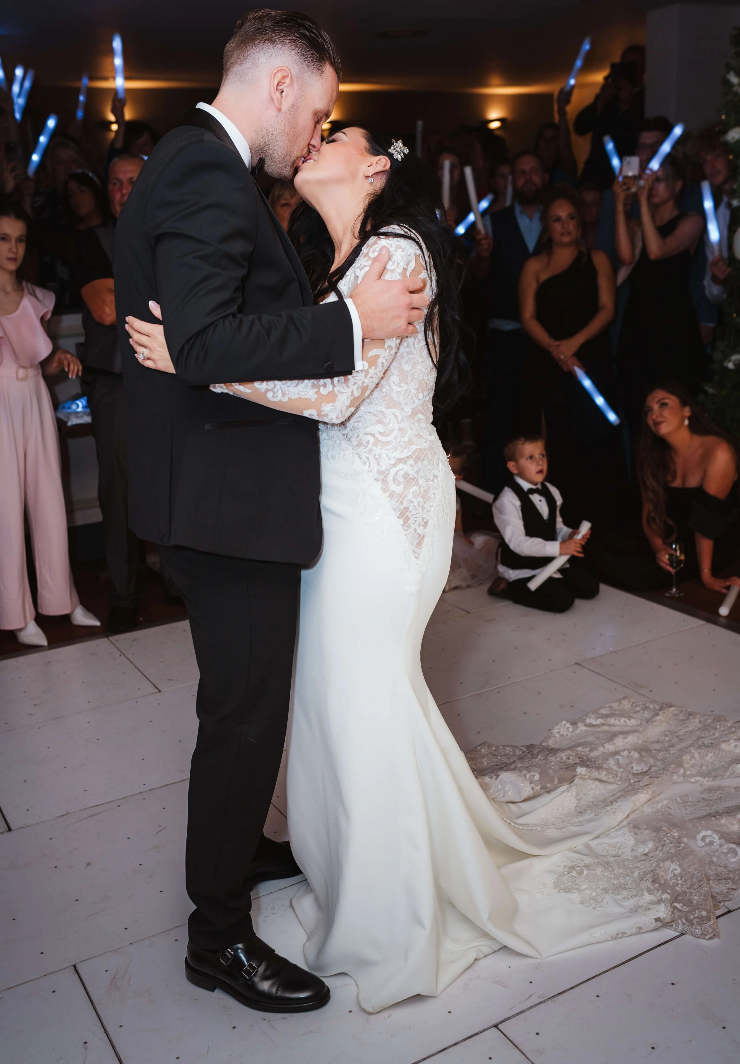 A newlywed couple sharing a kiss during their wedding reception, surrounded by wedding guests holding glow sticks.