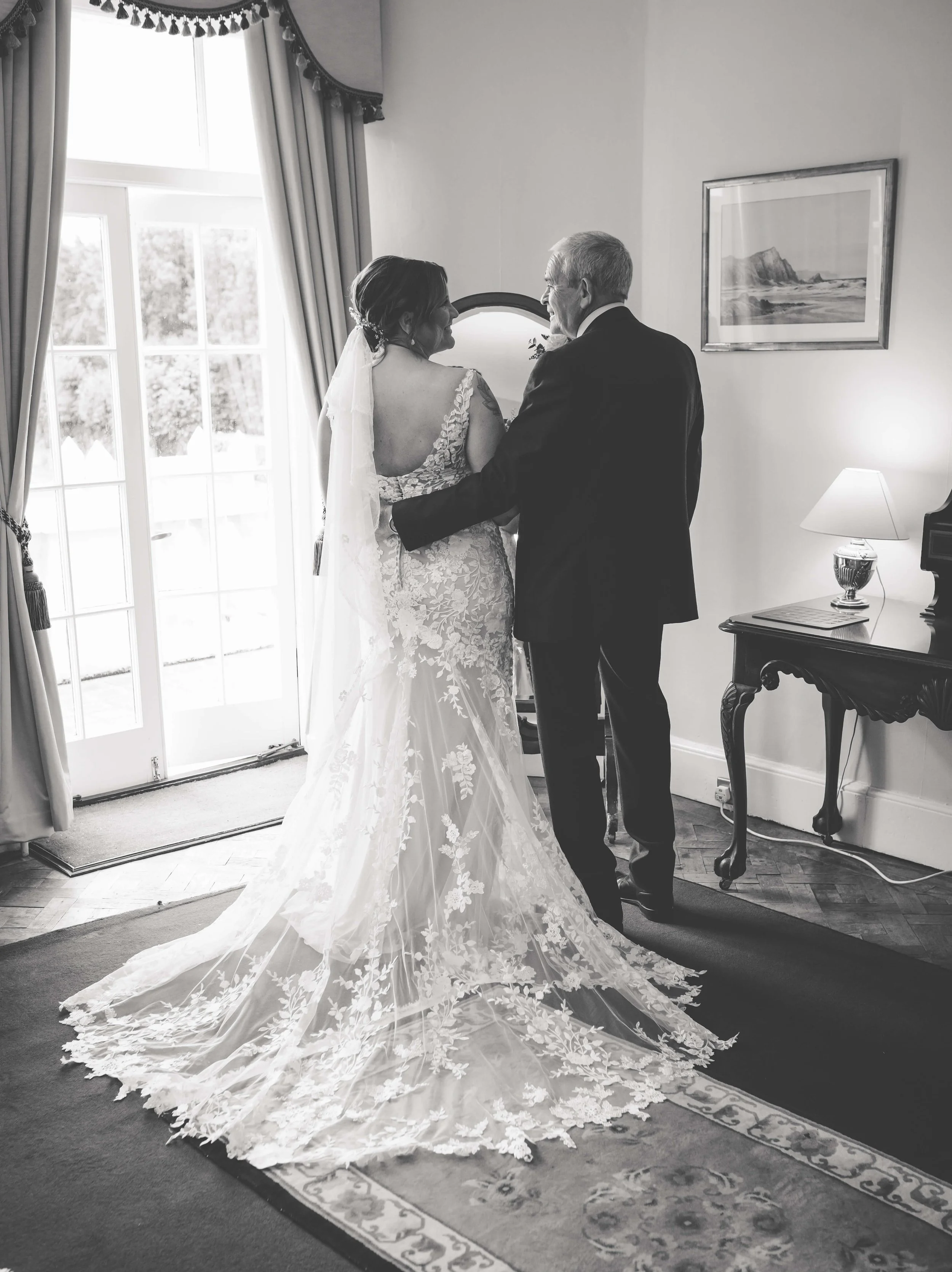 Black and white photo of a bride and an older man, possibly her father, standing in a room with a window and curtains. The bride is wearing a lace wedding gown with a long train and veil, and the man is in a suit. They are facing each other, holding 