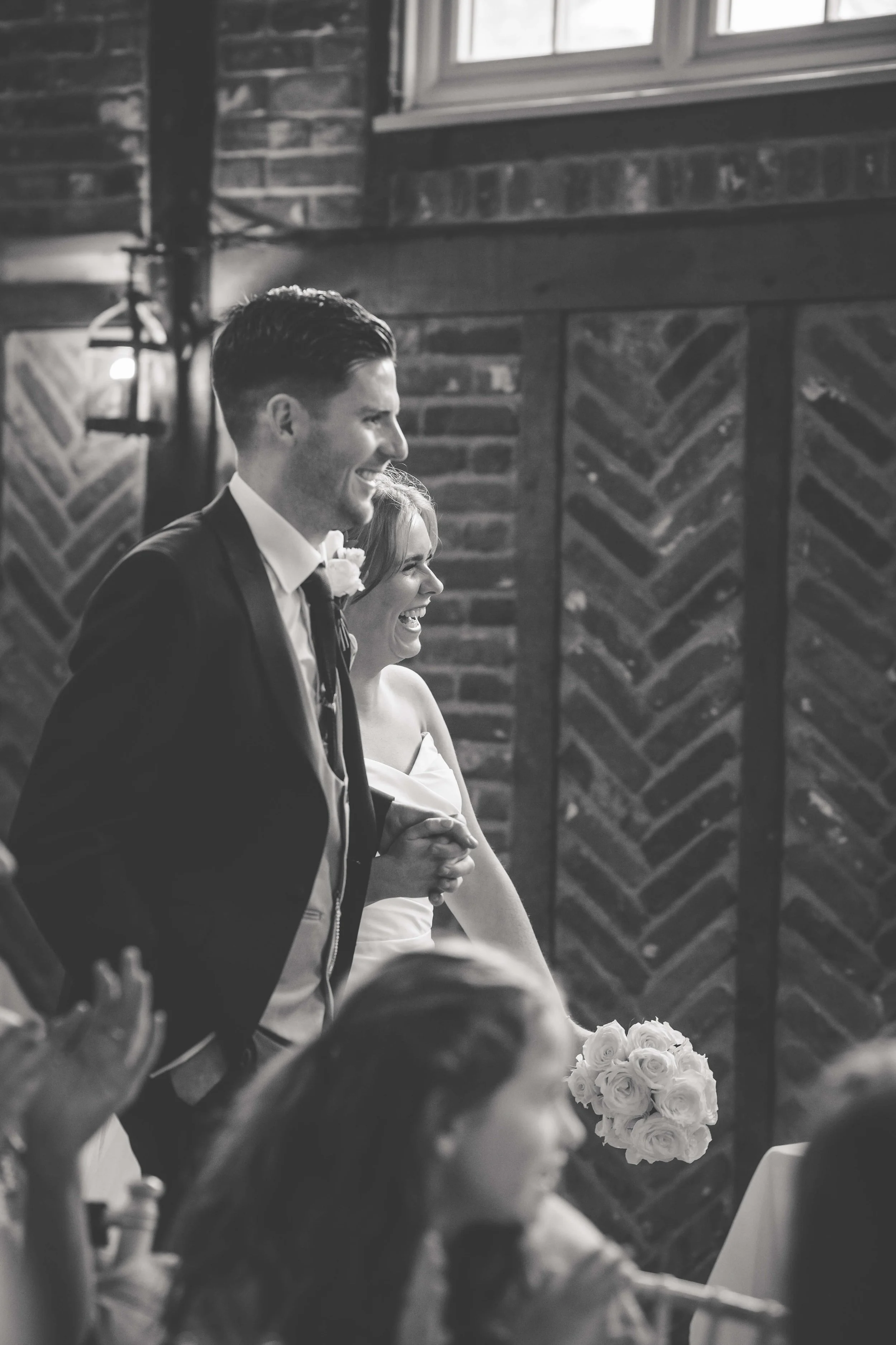 A black and white photo of a man and woman smiling during a wedding ceremony. The man is dressed in a suit and the woman in a wedding dress holding a bouquet of flowers. They are holding hands and seem happy, with a brick wall background and several 