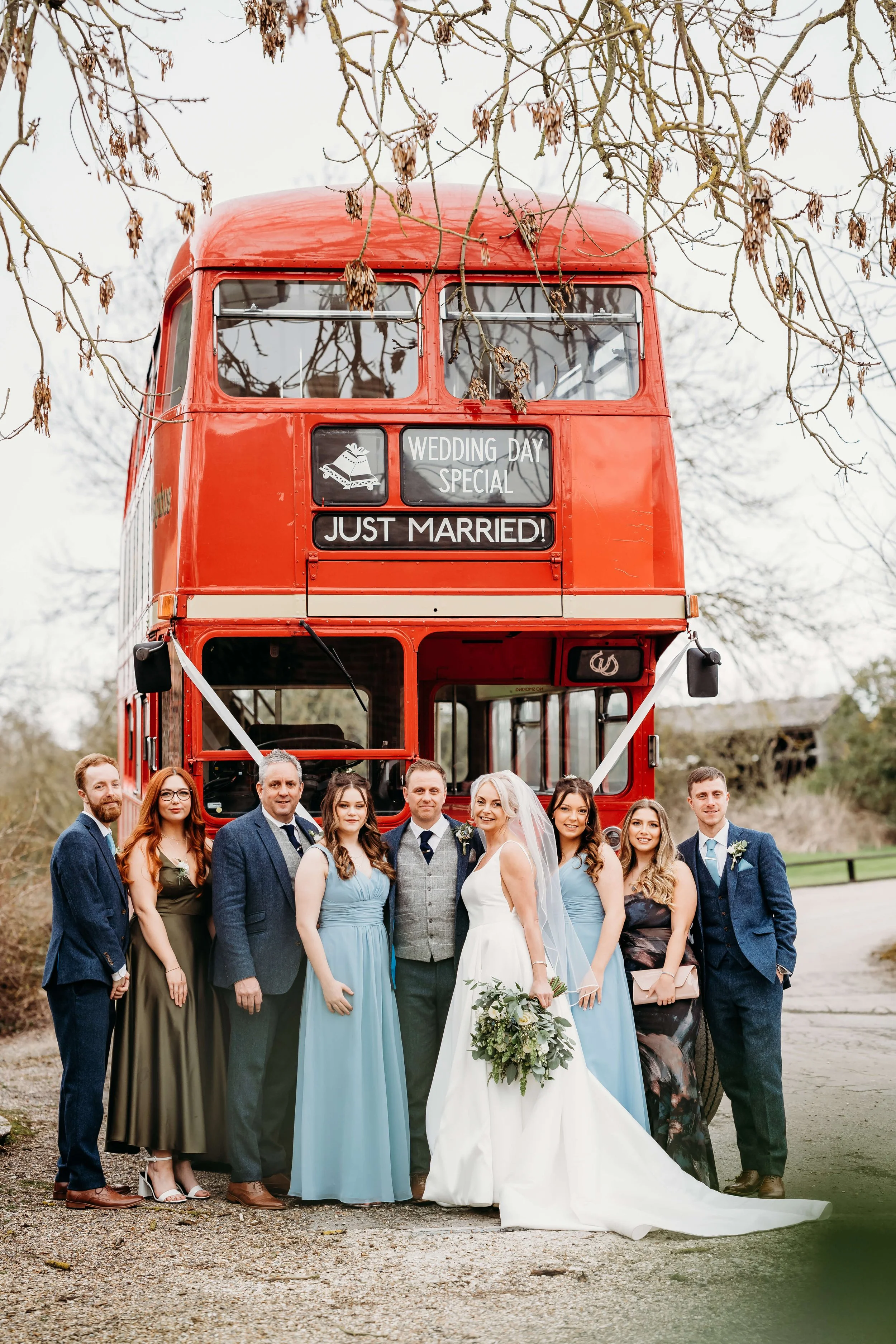 Wedding party standing in front of a red double-decker bus decorated for a wedding, with signs that say 'Wedding Day Special' and 'Just Married!'.