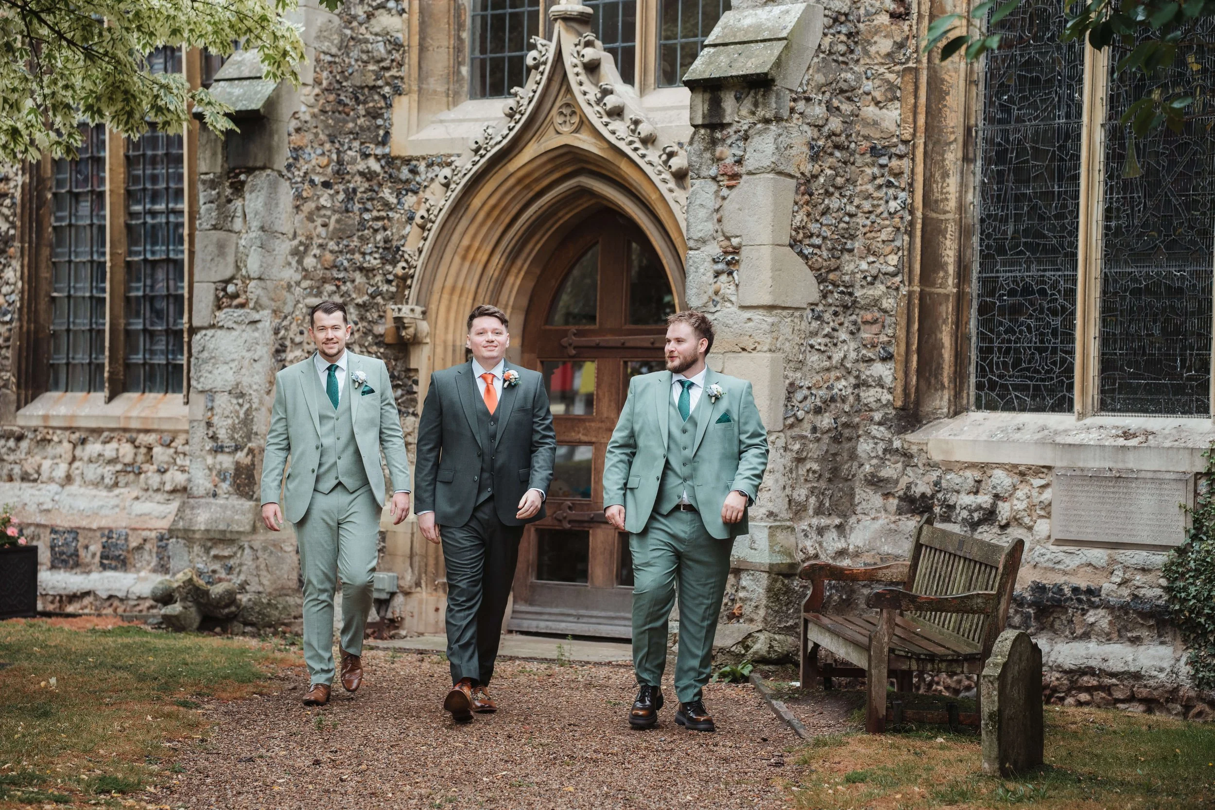 Three men in suits walking outside a historic stone building with stained glass windows and an arched doorway.