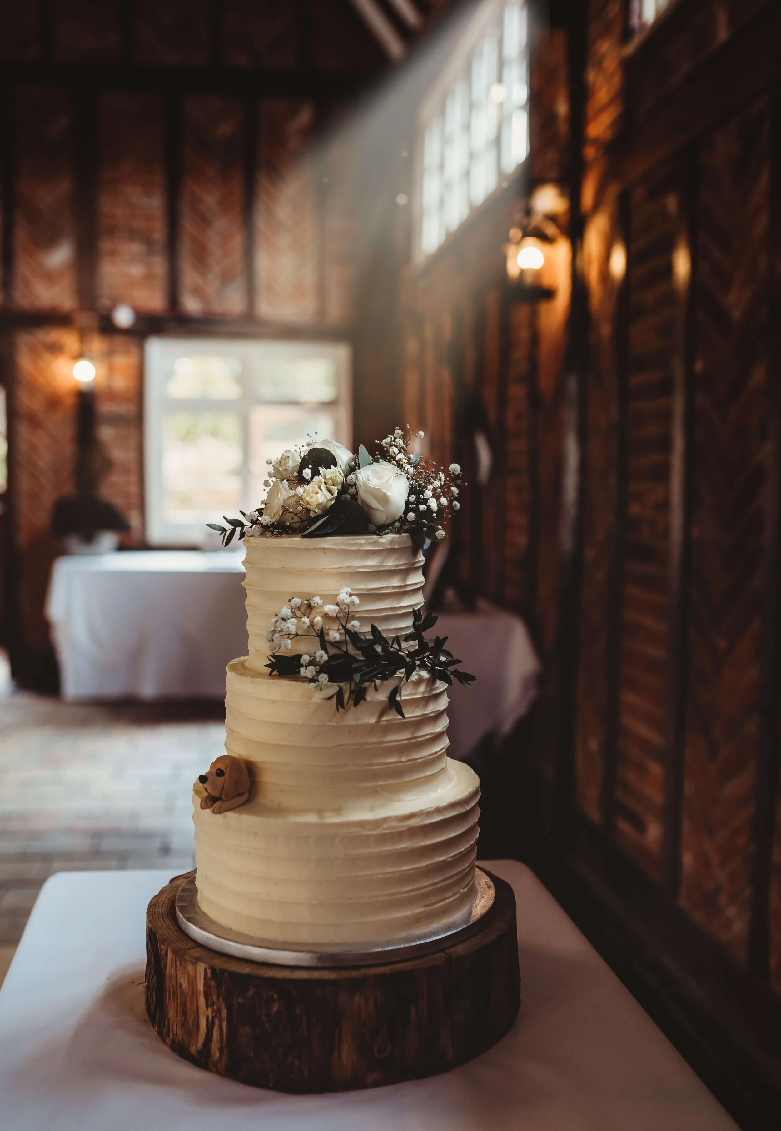 A three-tier white wedding cake with textured icing, decorated with white roses, baby's breath, and greenery on top. A small dog figurine is attached to the side of the cake. The cake is placed on a wooden slab on a white table in a rustic indoor set