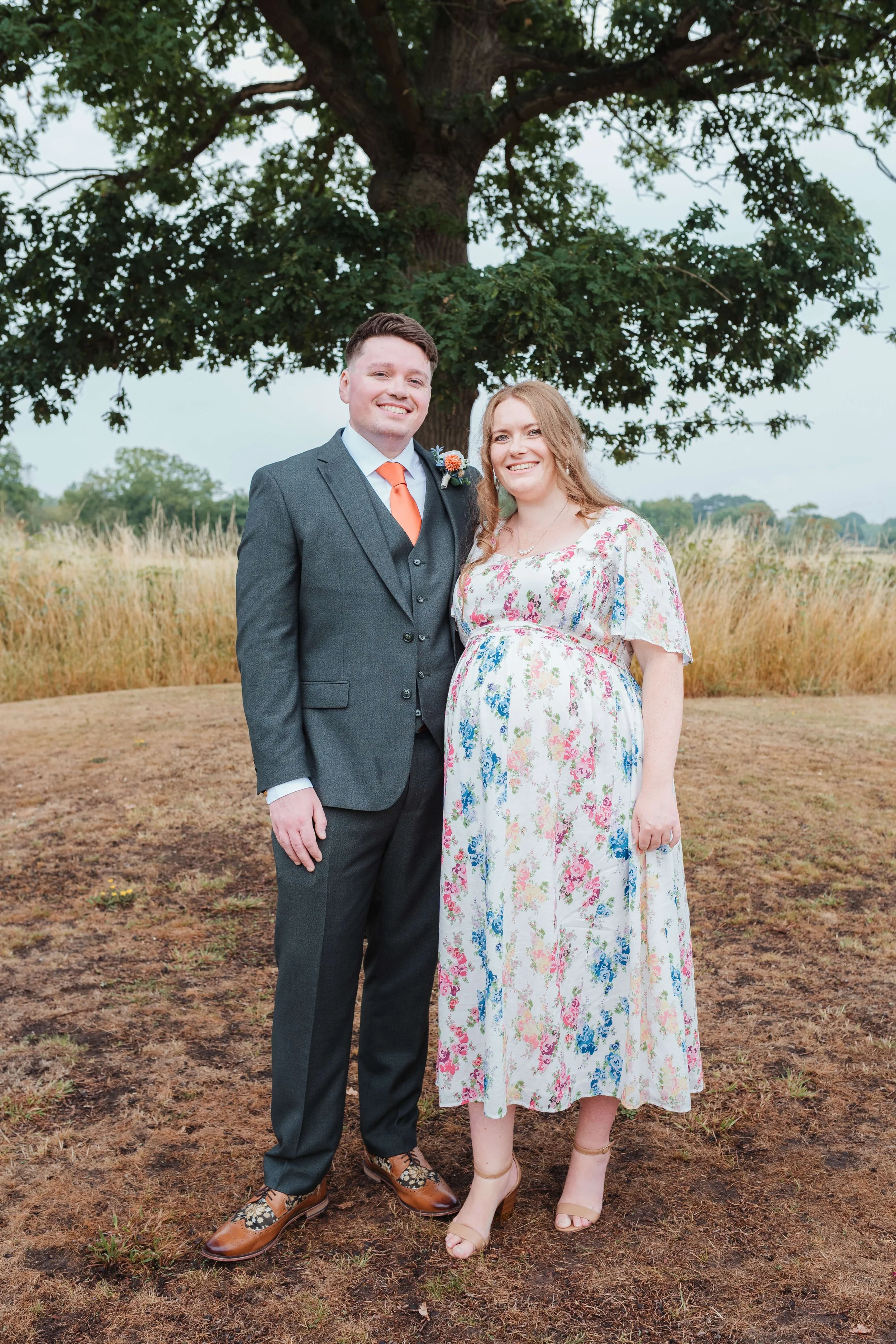 A couple standing outdoors, smiling, in front of a large tree with grassy field in the background. The man is dressed in a dark suit with an orange tie and boutonniere, and the woman is wearing a floral dress and high heels.