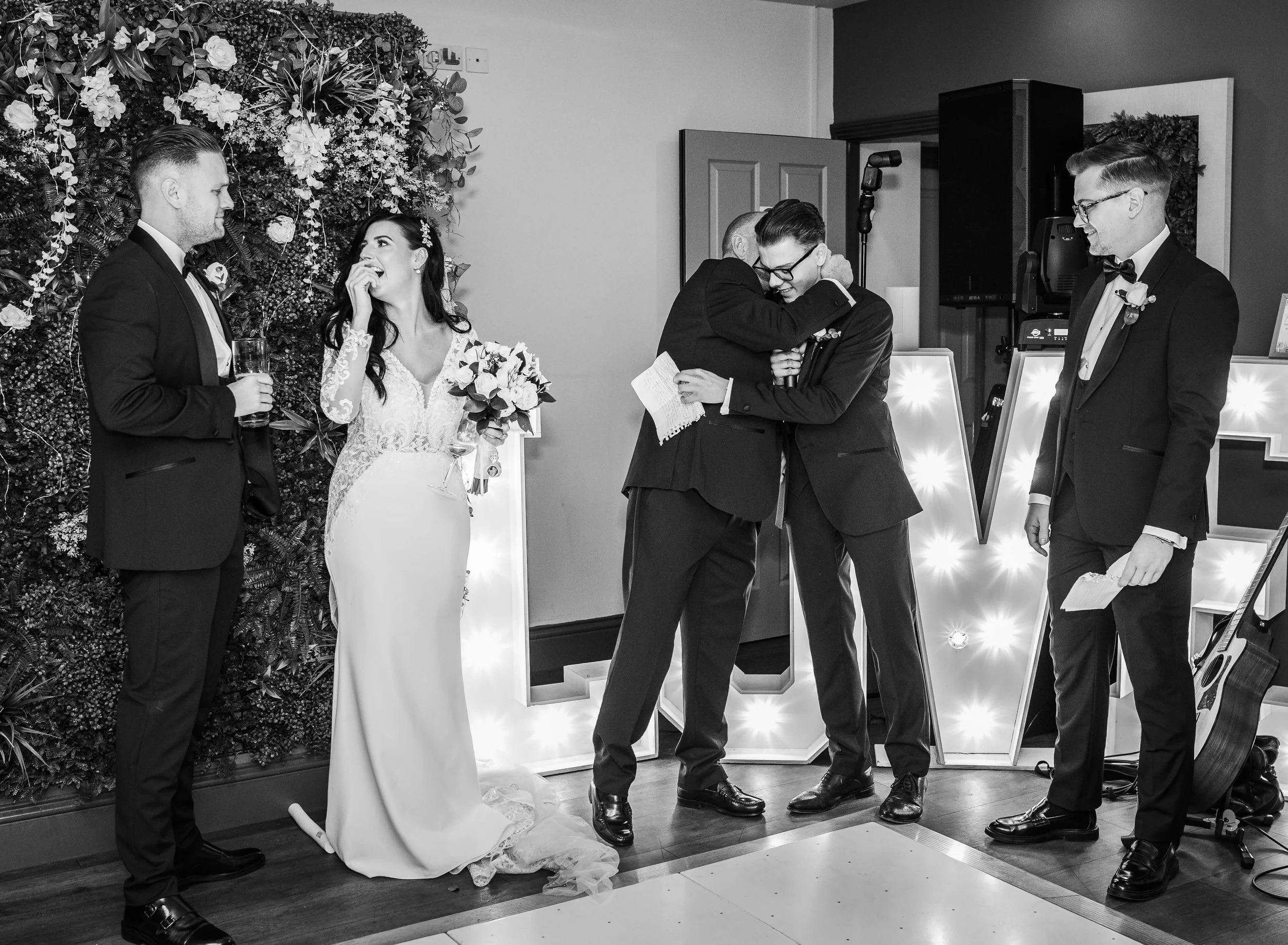 A black and white photo of a wedding reception. The bride and groom are hugging each other at the center. The bride is holding a bouquet of flowers, and the groom is holding a piece of paper. To the left, a woman in a wedding dress is smiling and loo