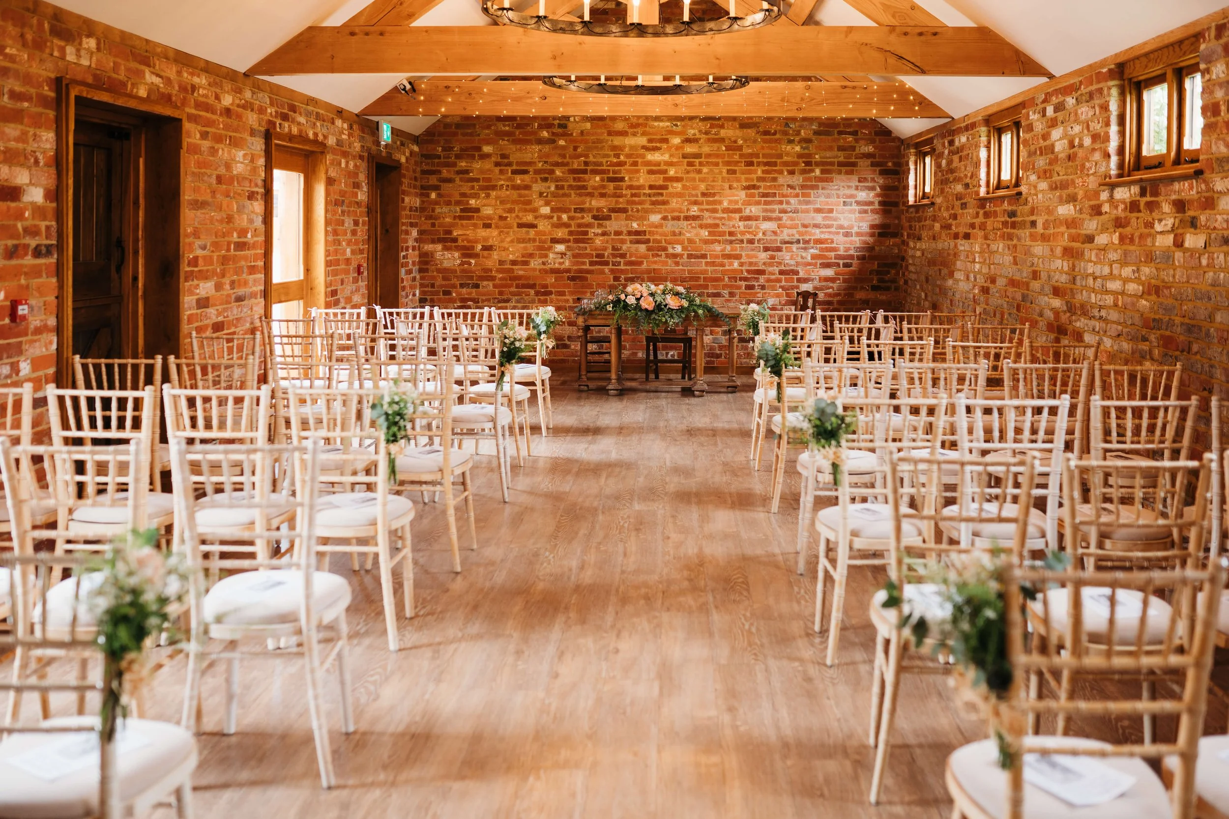 An indoor wedding ceremony setup with rows of white chairs decorated with small flower arrangements. The chairs face a small table with a floral centerpiece against a brick wall background and a wooden ceiling with string lights.