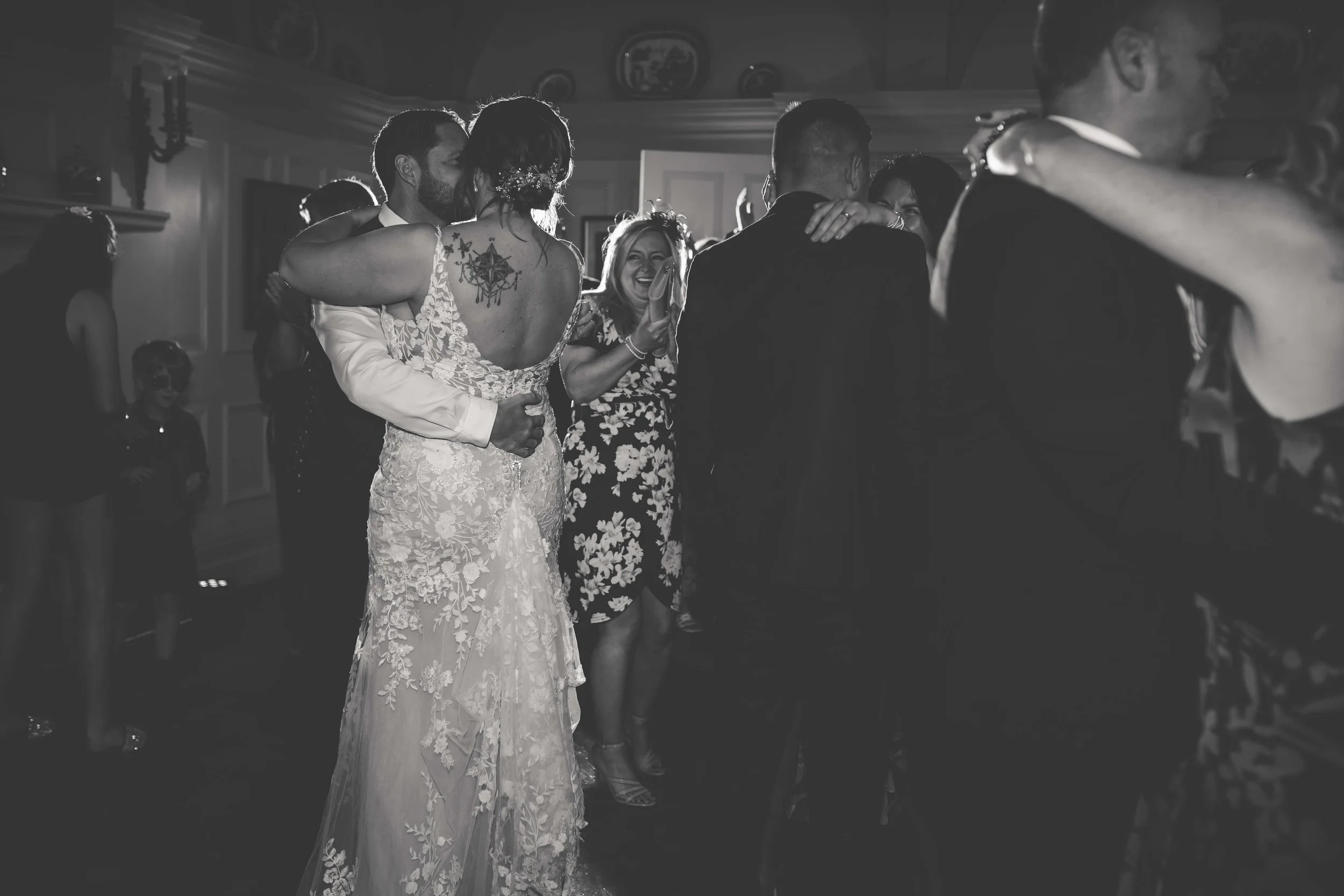 Black and white photo of a wedding reception with the bride and groom dancing closely while smiling, surrounded by guests in a decorated indoor venue.