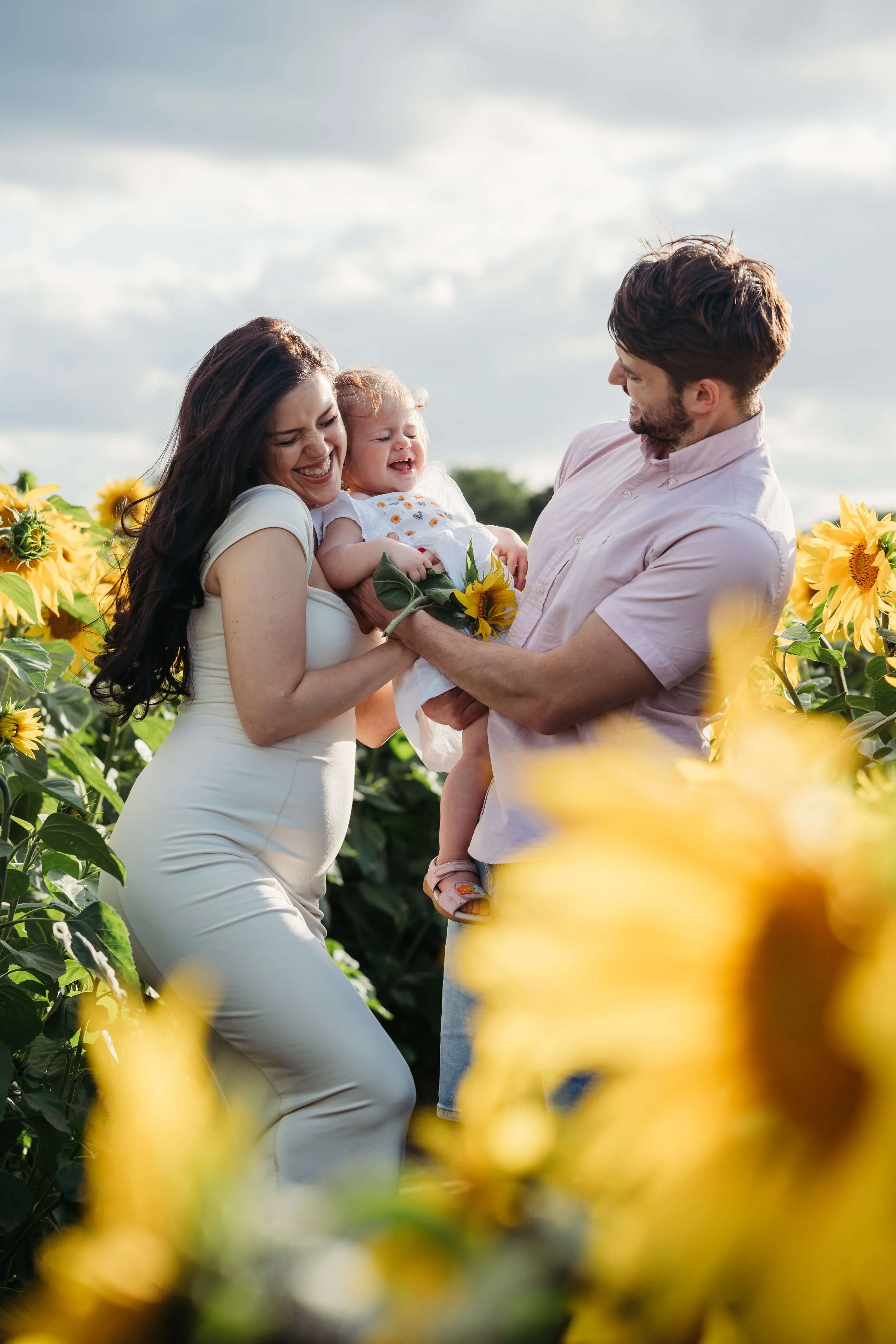 A family of three, a mother, father, and young girl, enjoying a moment together in a sunflower field. The mother, dressed in white, is holding her daughter, who is laughing, while the father stands facing them with a smile, holding a sunflower.