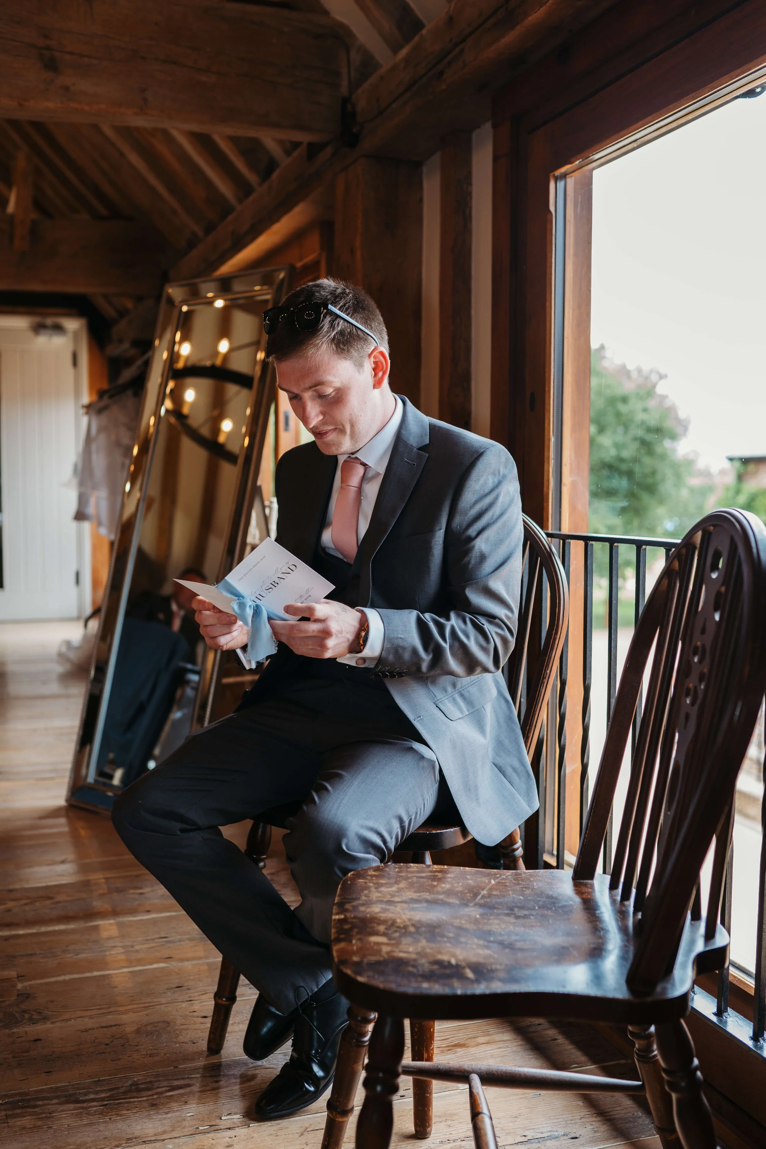 A young man in a suit sitting on a wooden chair, reading a card near a large window inside a rustic cabin.