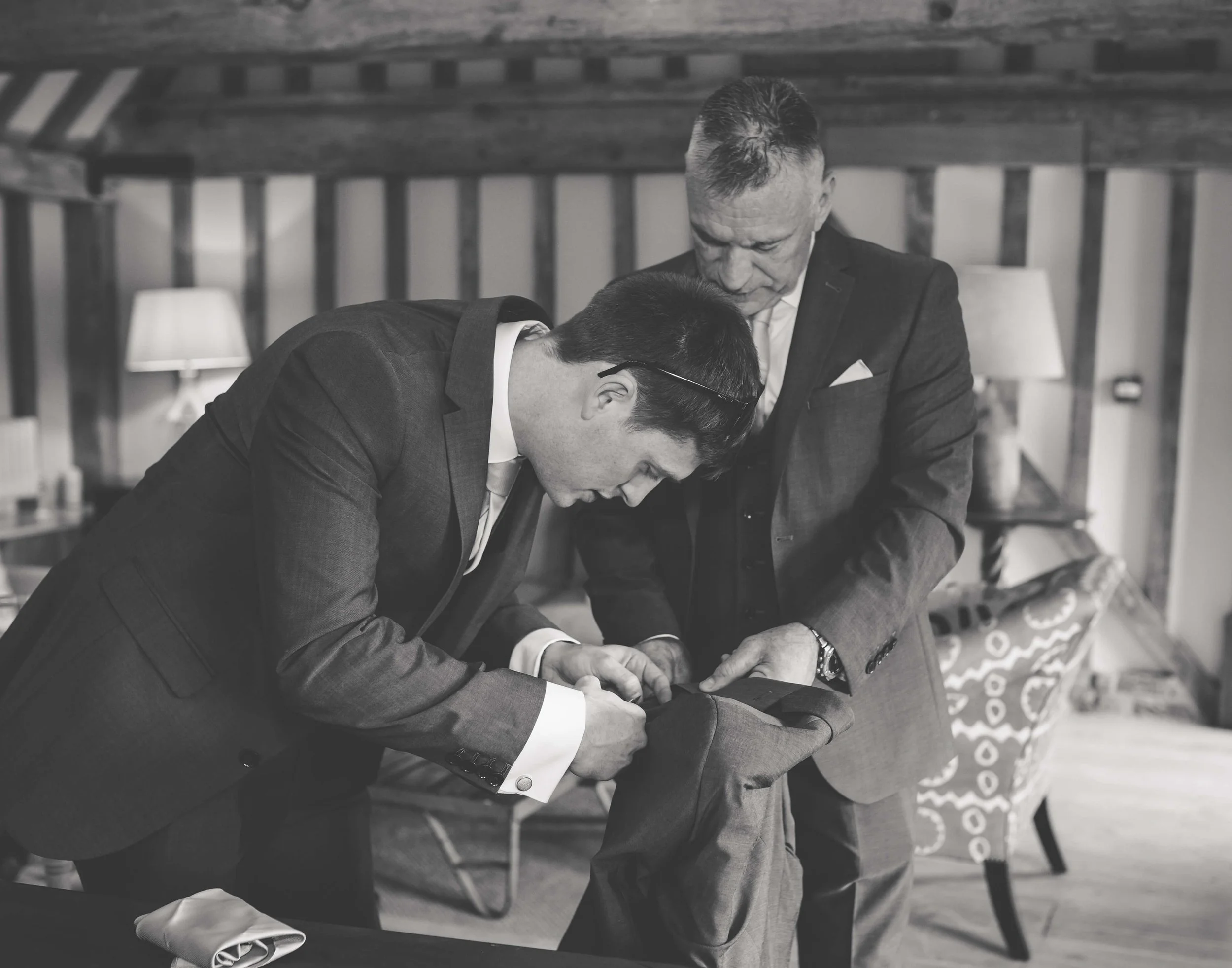 Two men in suits adjusting the cuff of a third man in a suit, in a room with wooden beams and a patterned chair.