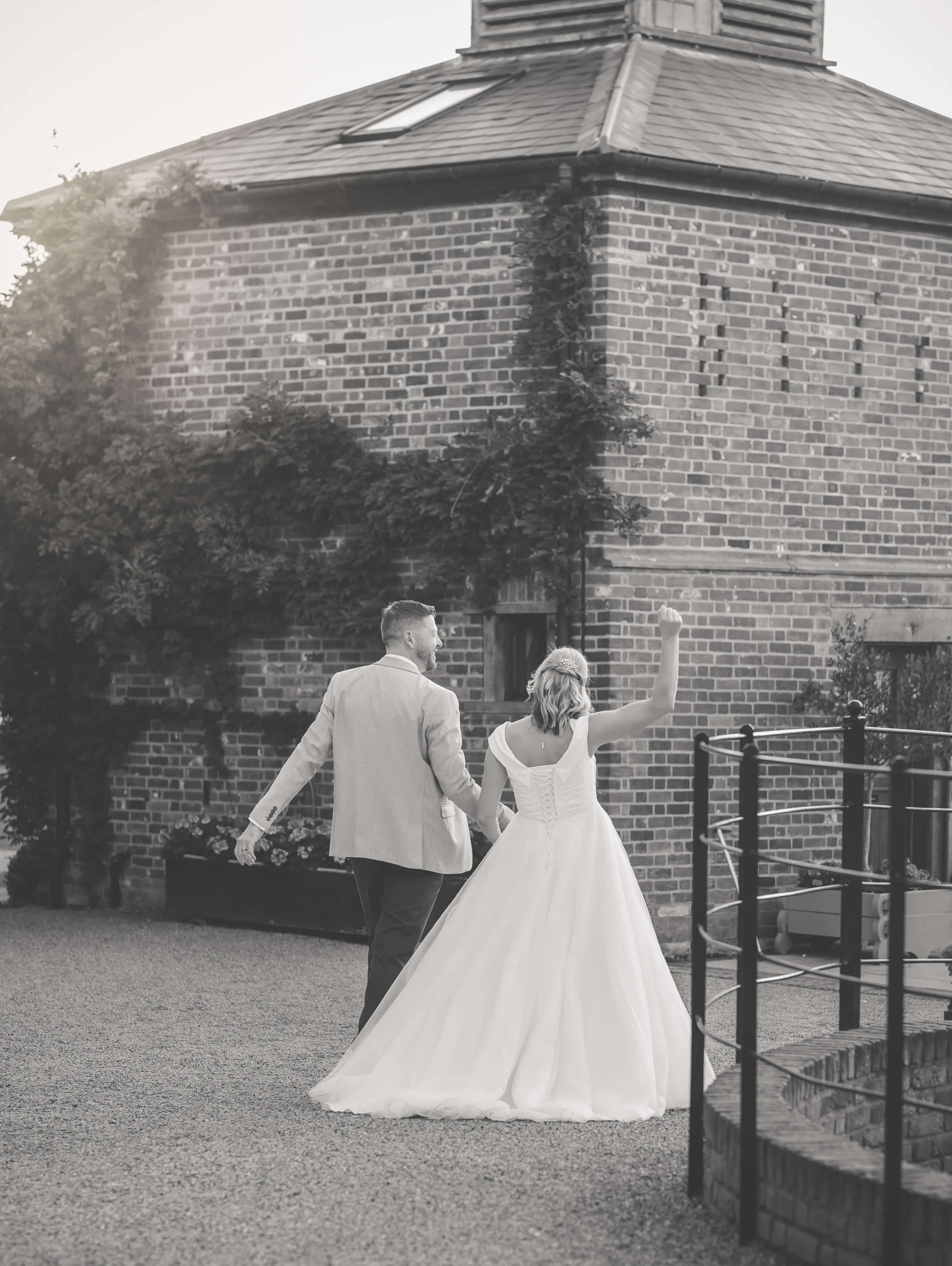 A bride and groom walking hand in hand outside a brick building, with the bride raising her fist in celebration.