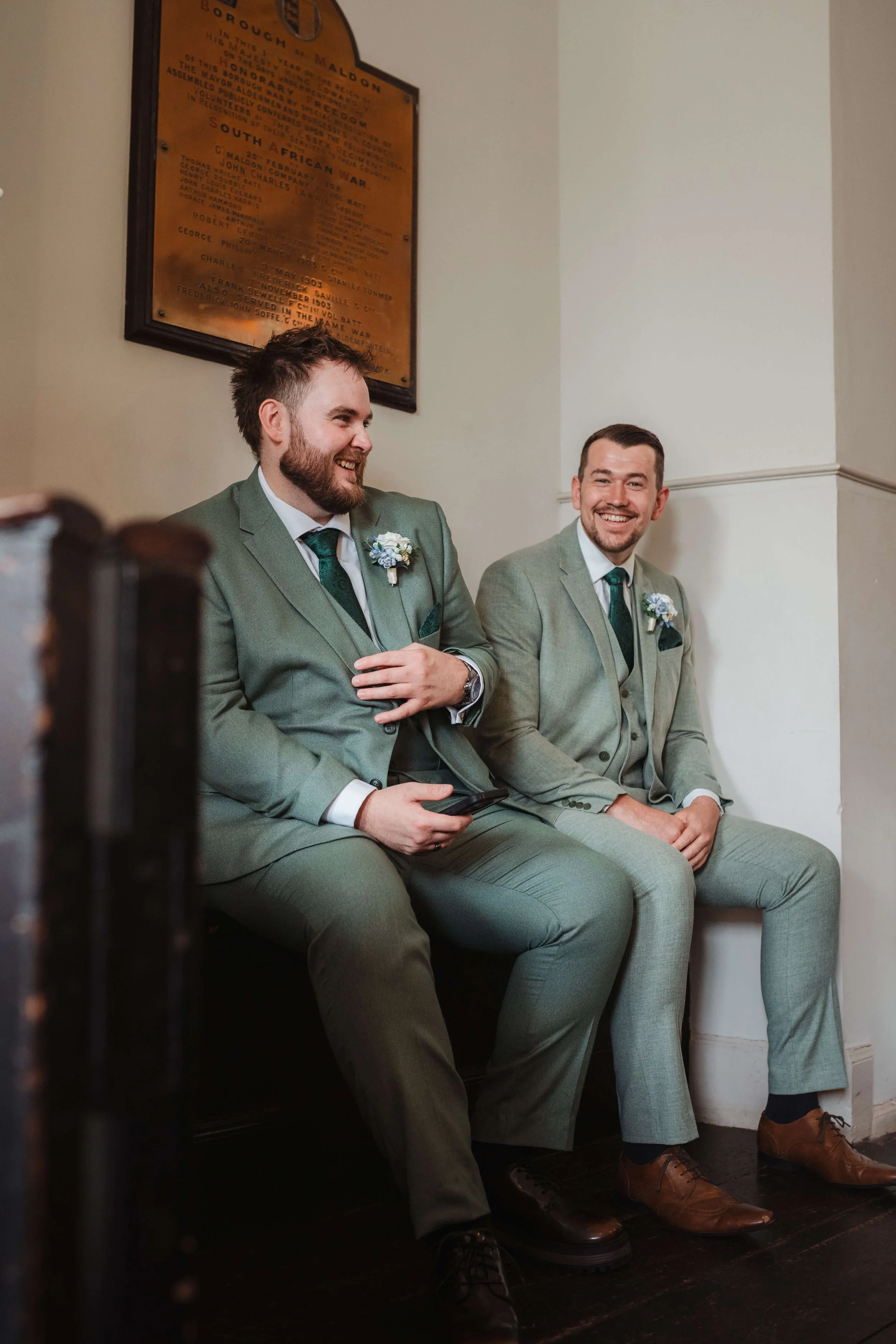 Two men in matching light green suits sitting on a bench, smiling and talking in a room with a framed historical plaque on the wall behind them.