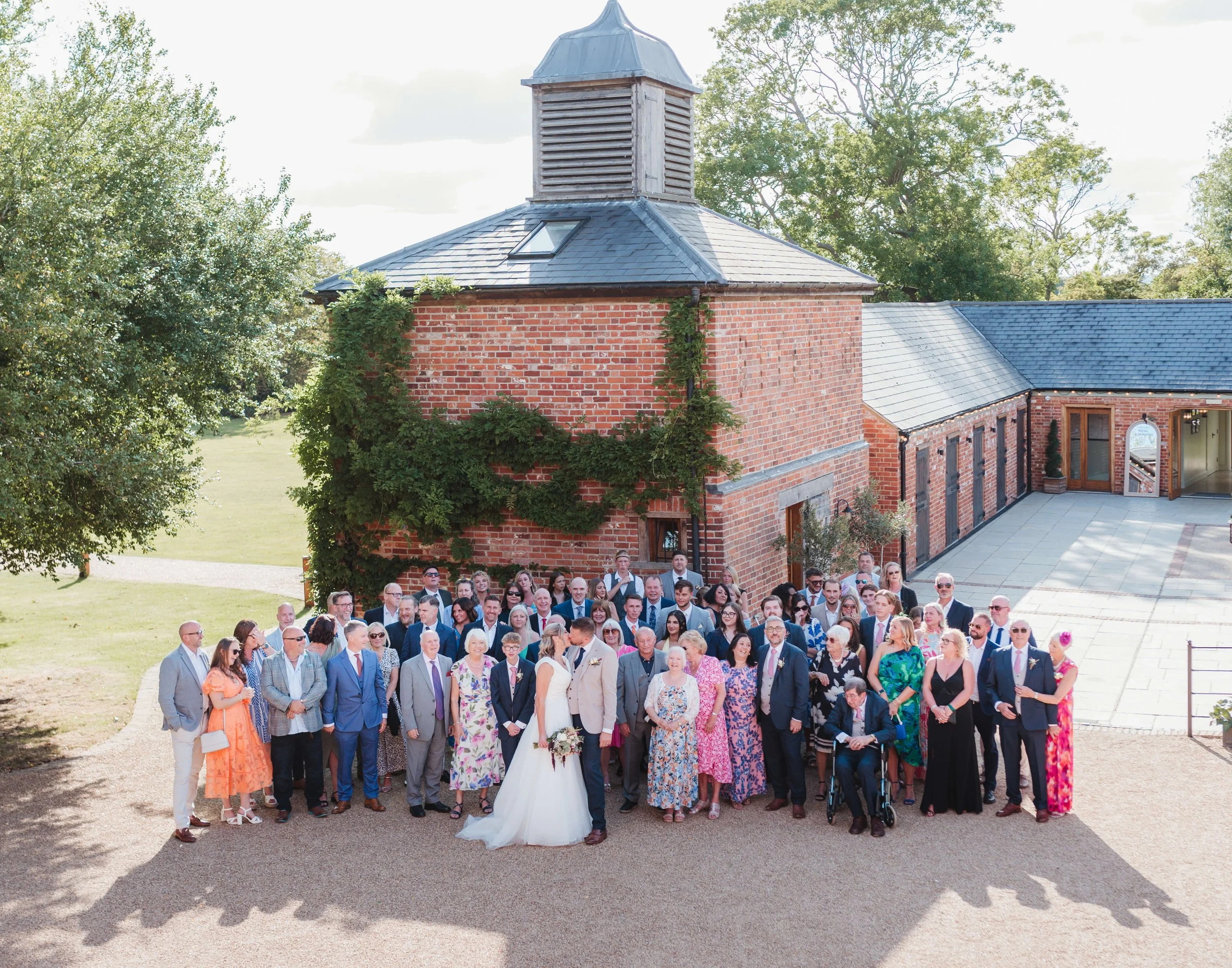 A large group of people, including a bride and groom, gathers outside a brick building and a church for a wedding photo on a sunny day.