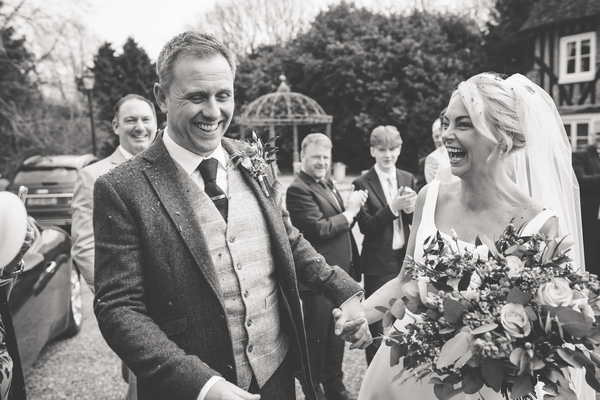 A black and white photo of a wedding scene with a happy bride and groom holding hands, surrounded by guests clapping and smiling outdoors.