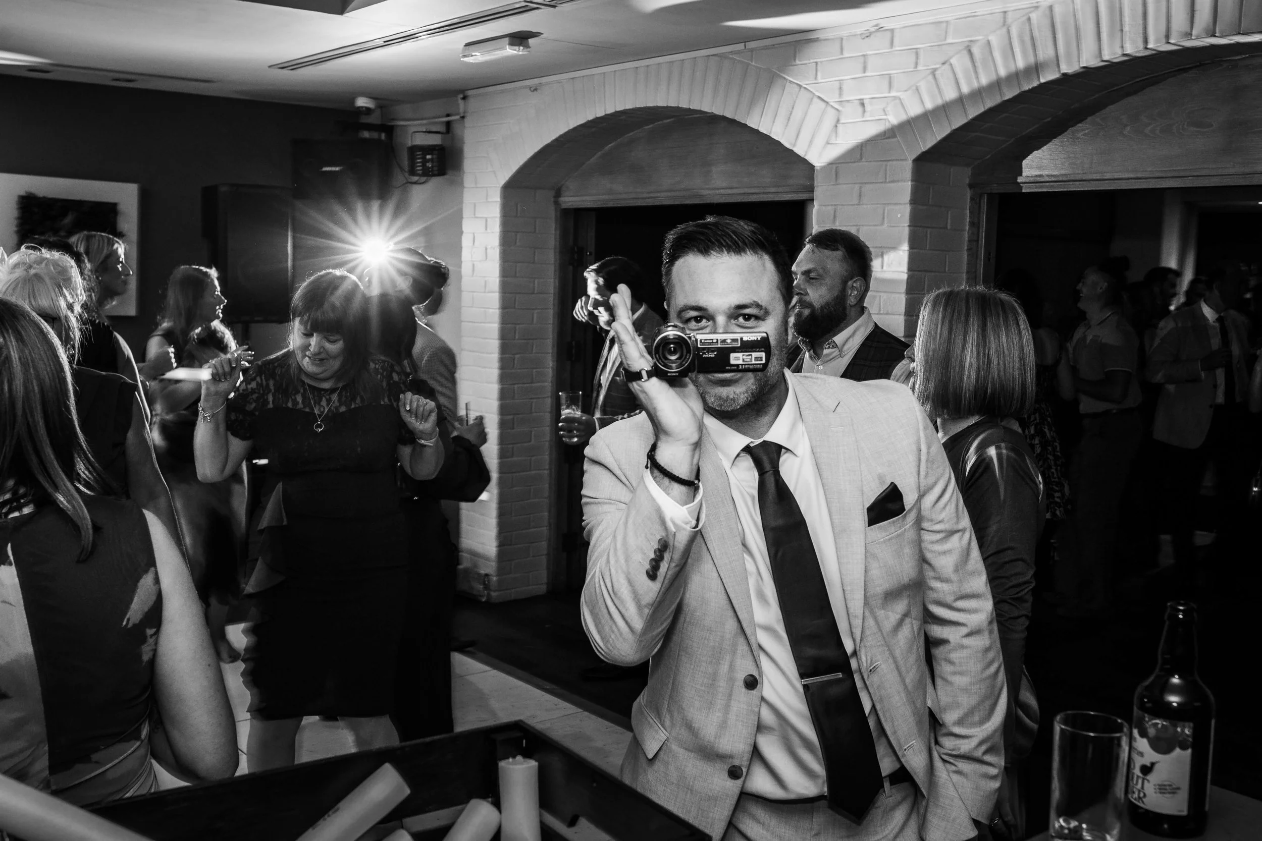 A man in a suit holding a video camera up to his face at a party, with guests dancing and socializing in the background, some with drinks in hand.