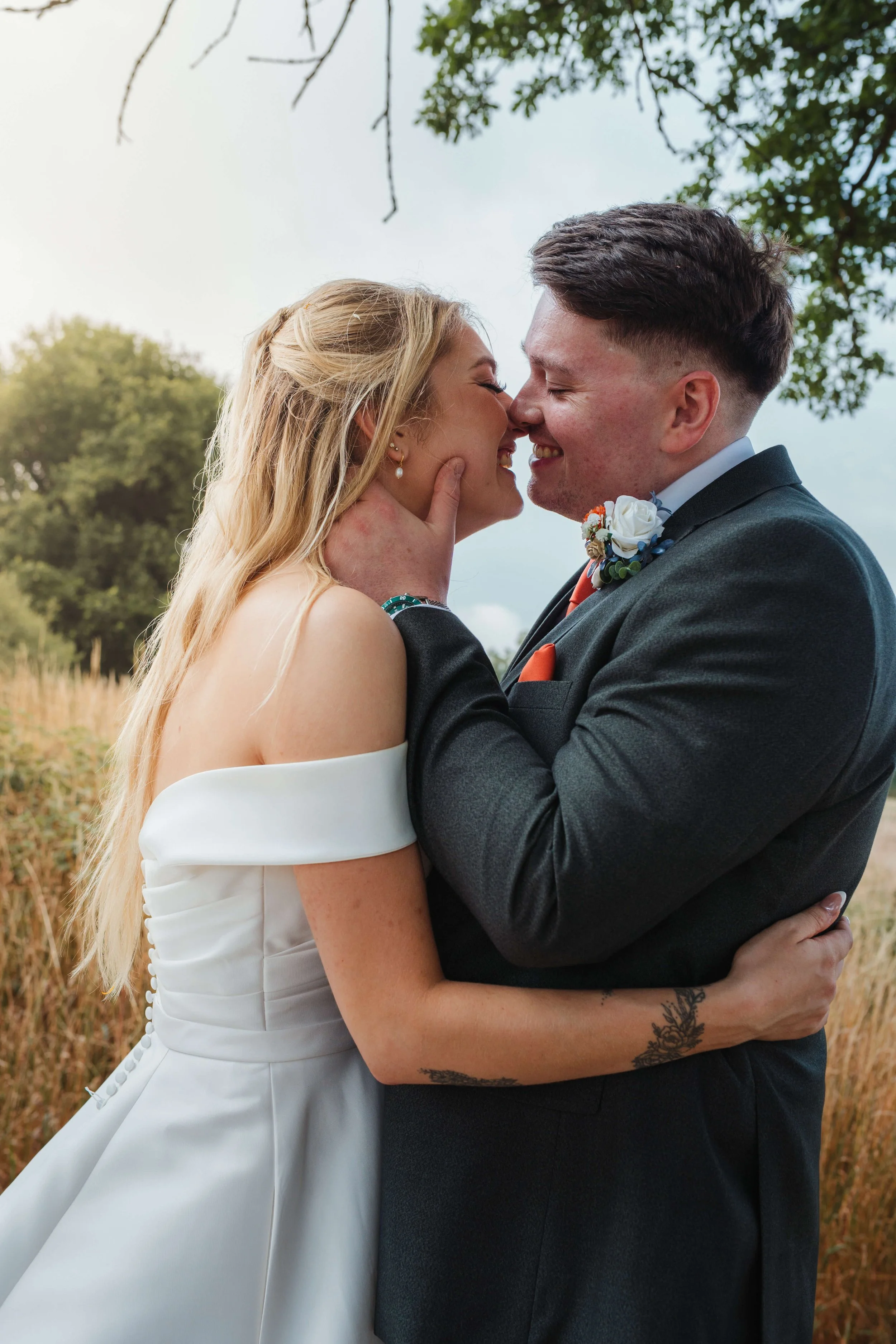 A bride and groom embrace outdoors, the bride in an off-shoulder white wedding dress and the groom in a gray suit, smiling with their noses touching.