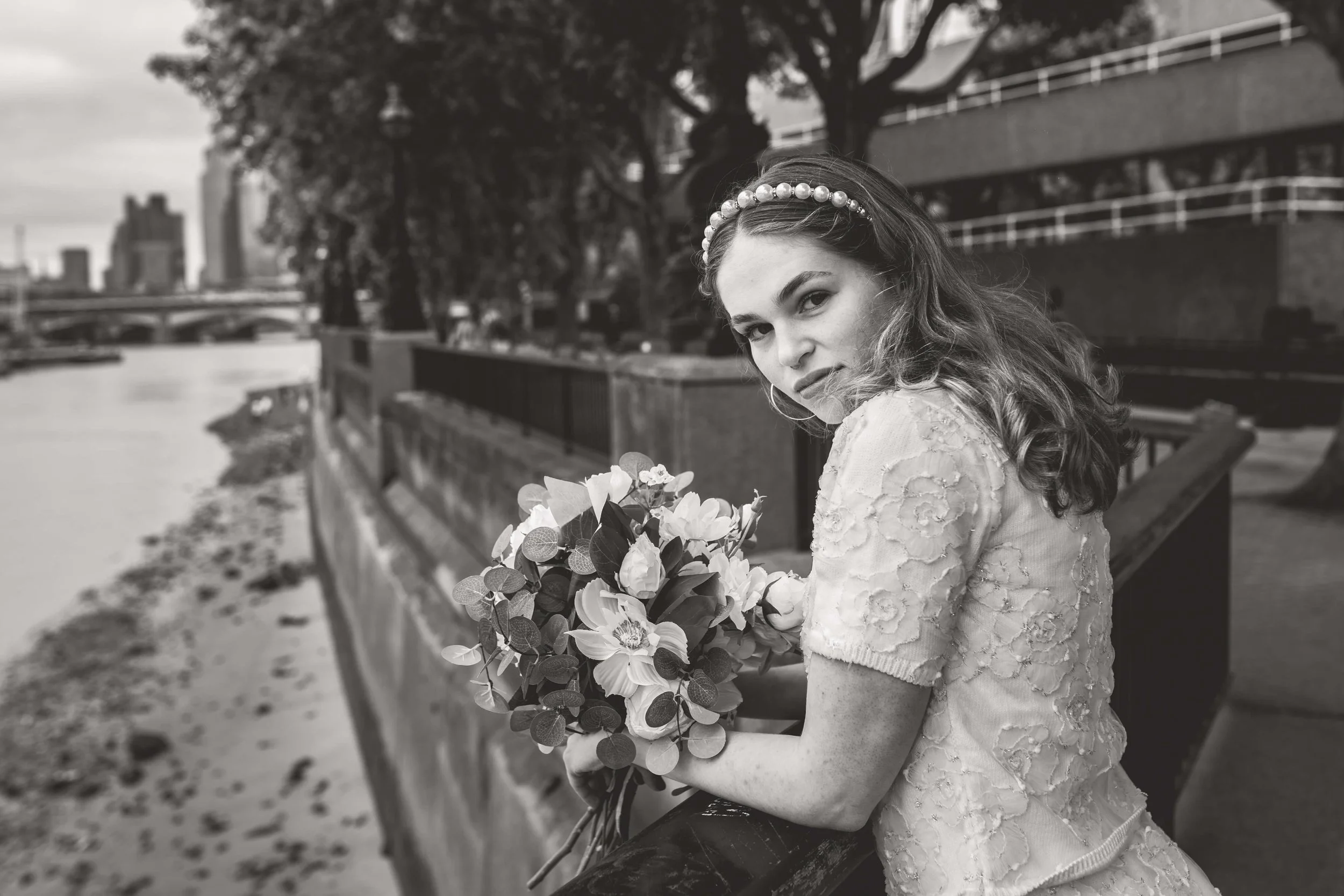 A black and white photograph of a young woman sitting on a wooden railing by a river, holding a bouquet of flowers, looking over her shoulder at the camera. She is wearing a lace dress and a pearl headband, with trees and buildings in the background.