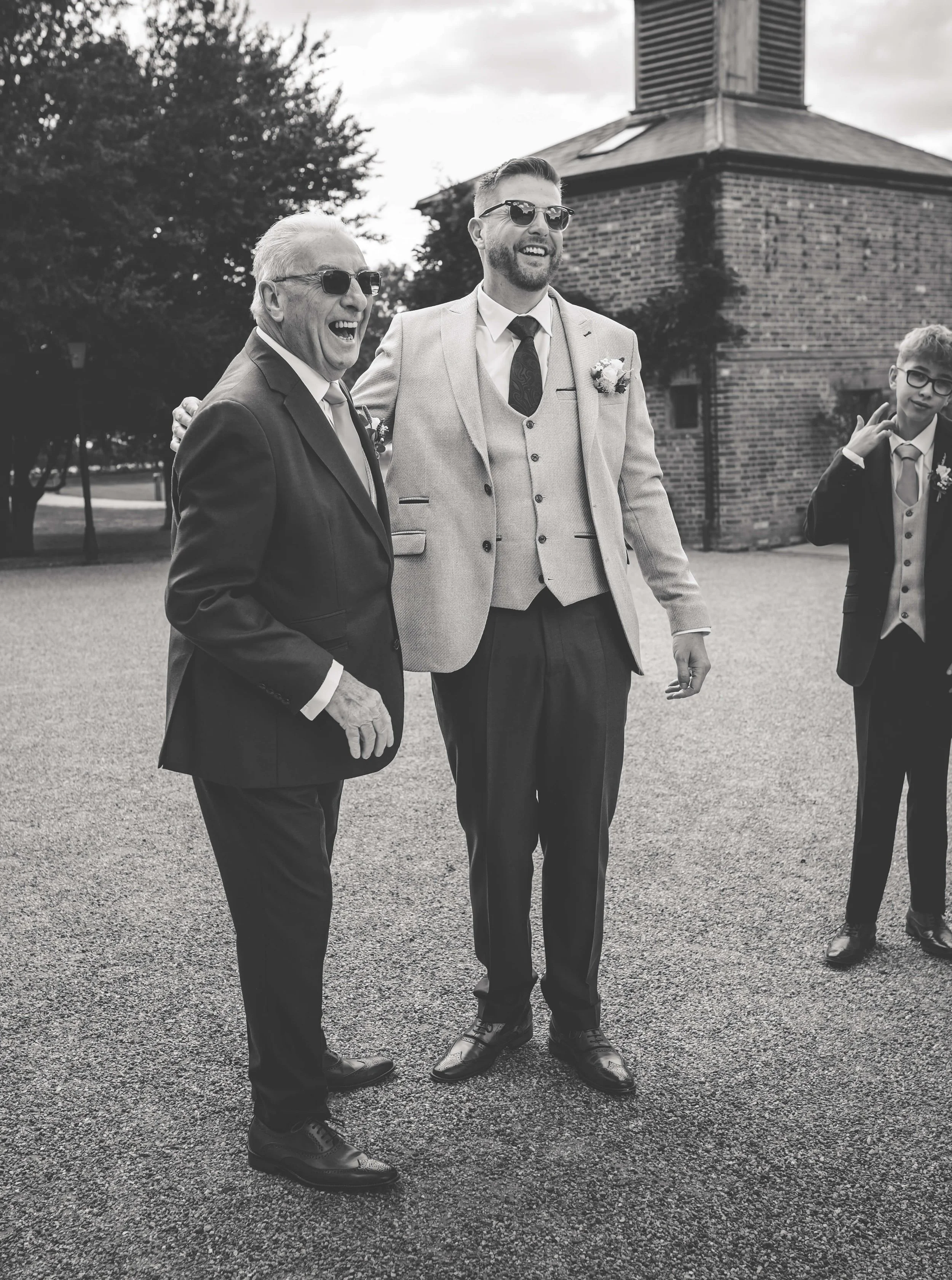 Black and white photograph of three males in suits, laughing and smiling outdoors, with a brick building and trees in the background.