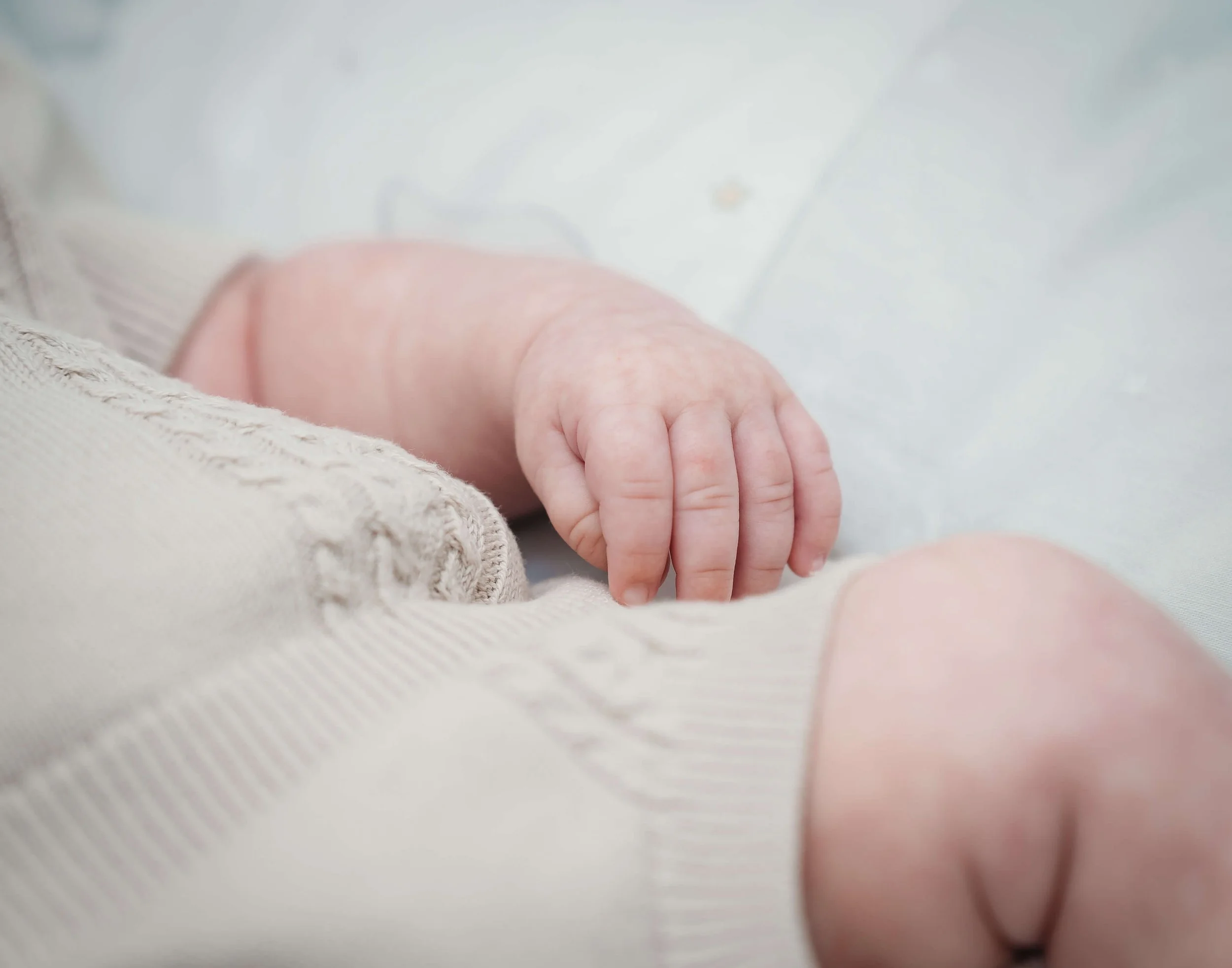 Close-up of a tiny baby's hand resting on a soft surface, wearing a beige or cream-colored outfit.