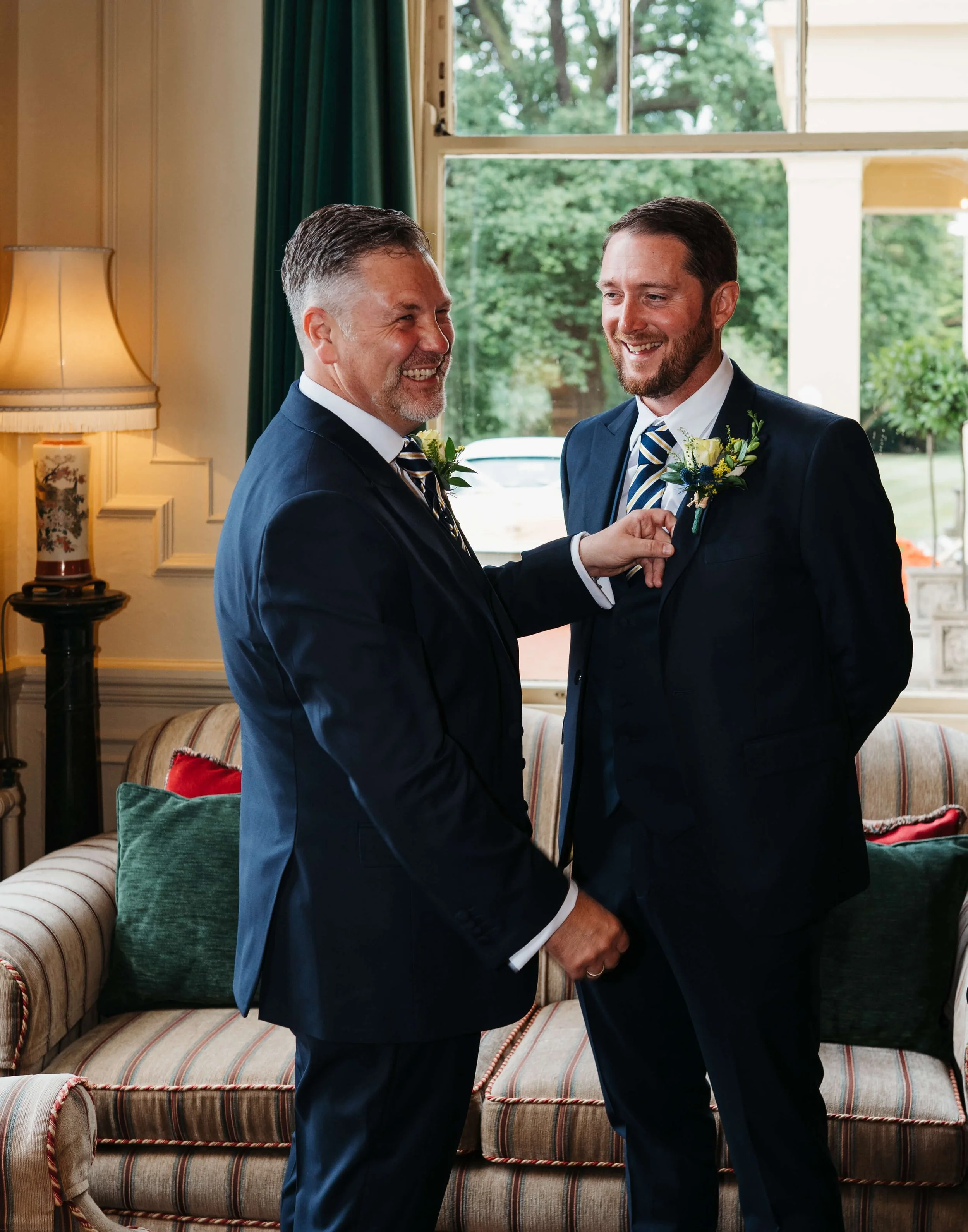 Two men dressed in navy suits and striped ties, smiling and sharing a moment indoors near a window with green trees outside. One man is adjusting his boutonniere thinned flower with his hand, the other man is holding the other man's hand. The room ha