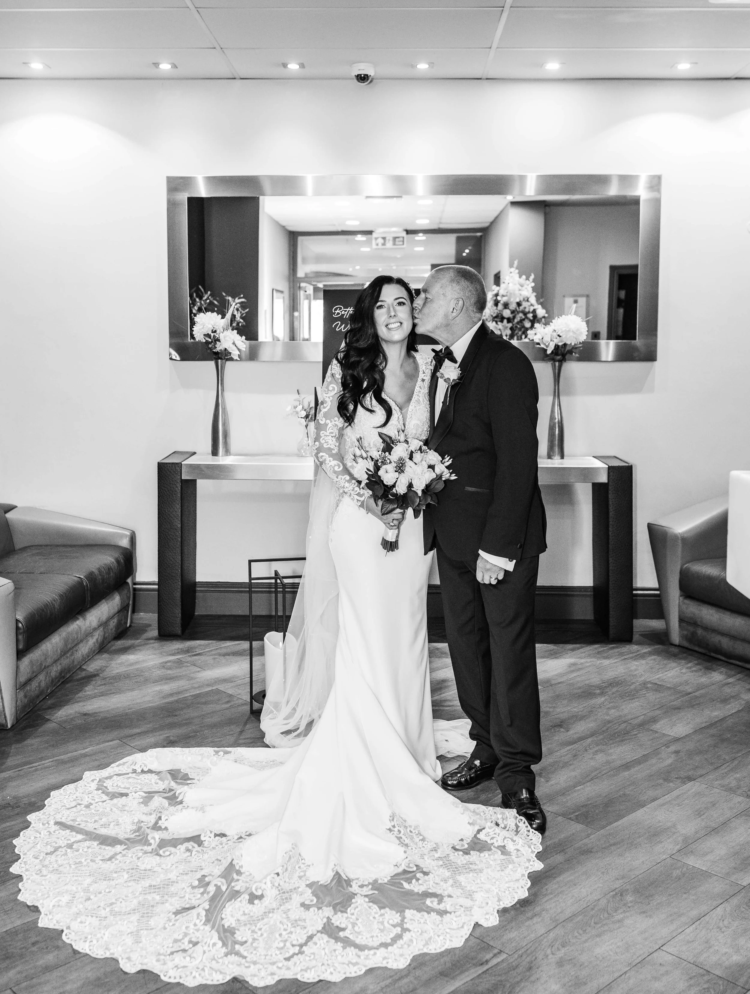 A bride and groom in wedding attire, with the groom kissing the bride on the cheek, standing in a modern indoor setting with decorations and a mirror reflecting the hallway.