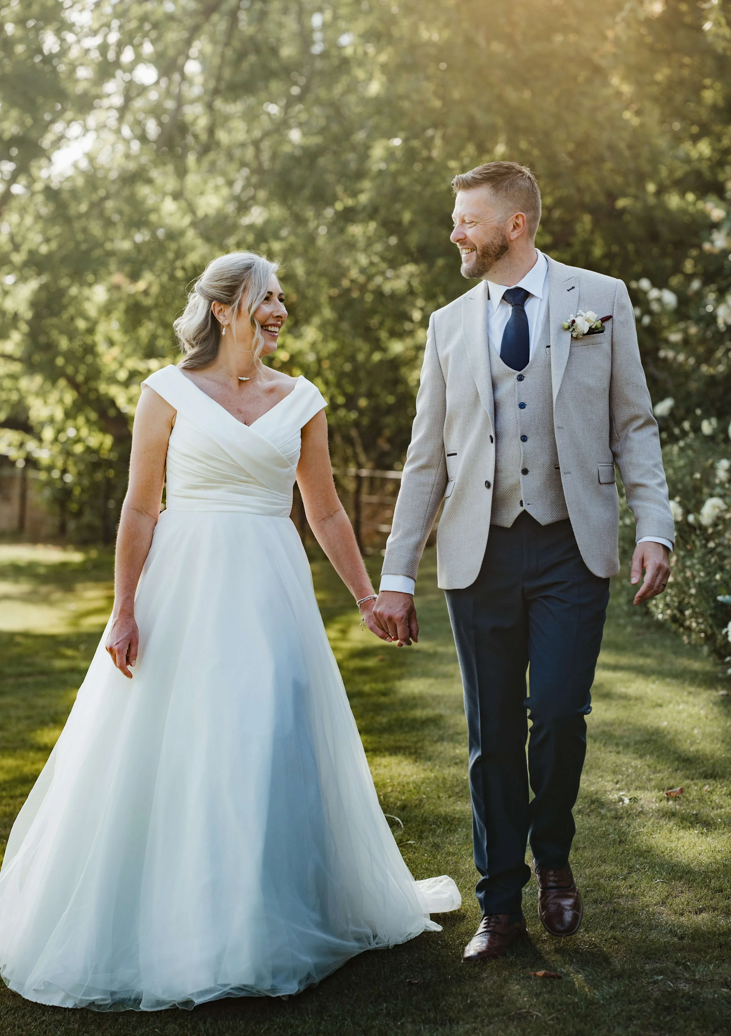 A bride and groom holding hands and smiling at each other outdoors in a garden during late afternoon or early evening.