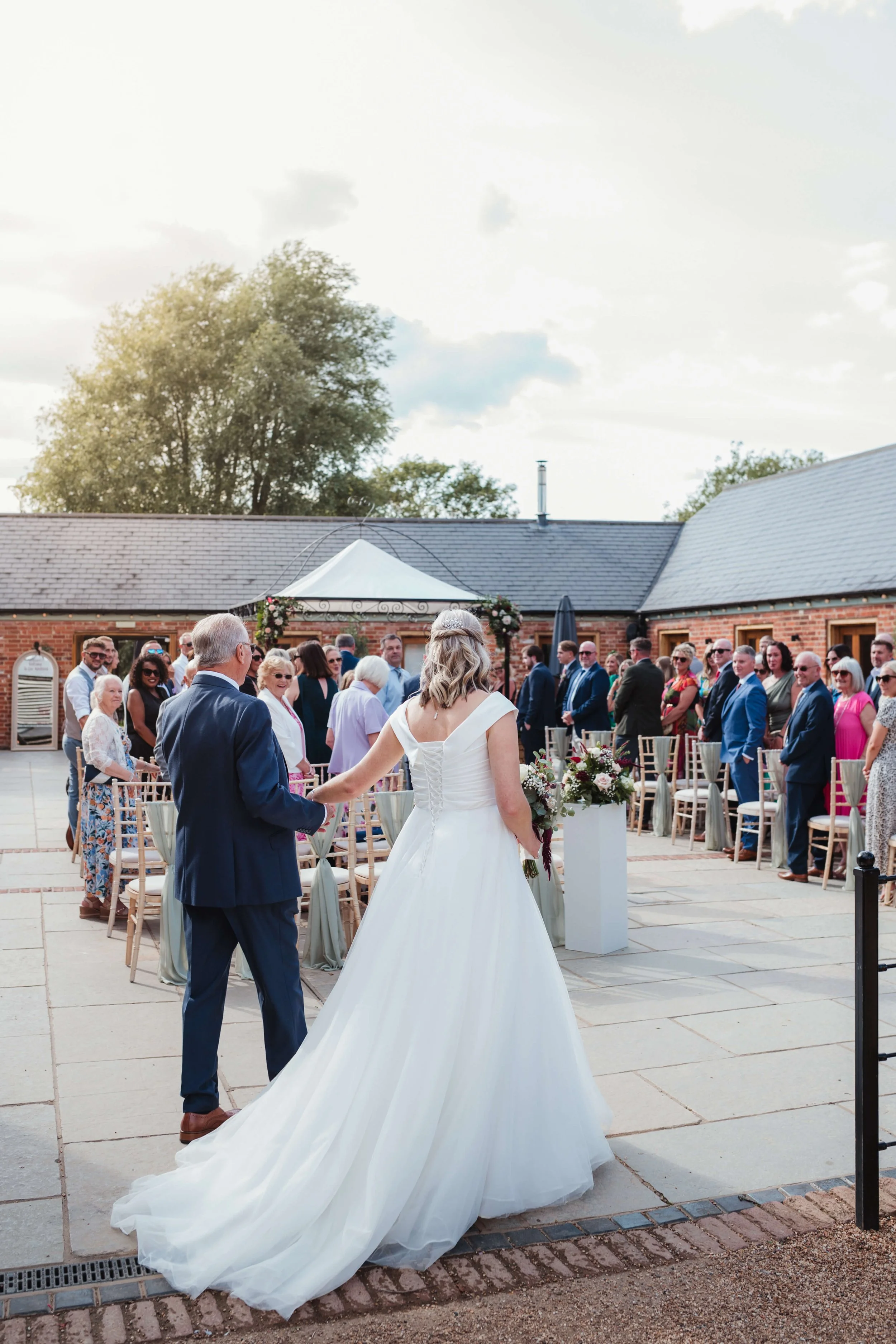 A bride in a white wedding dress holding hands with an elderly man, likely her father, at an outdoor wedding ceremony. Guests are standing and seated on either side, with a wedding arch decorated with flowers in the background.