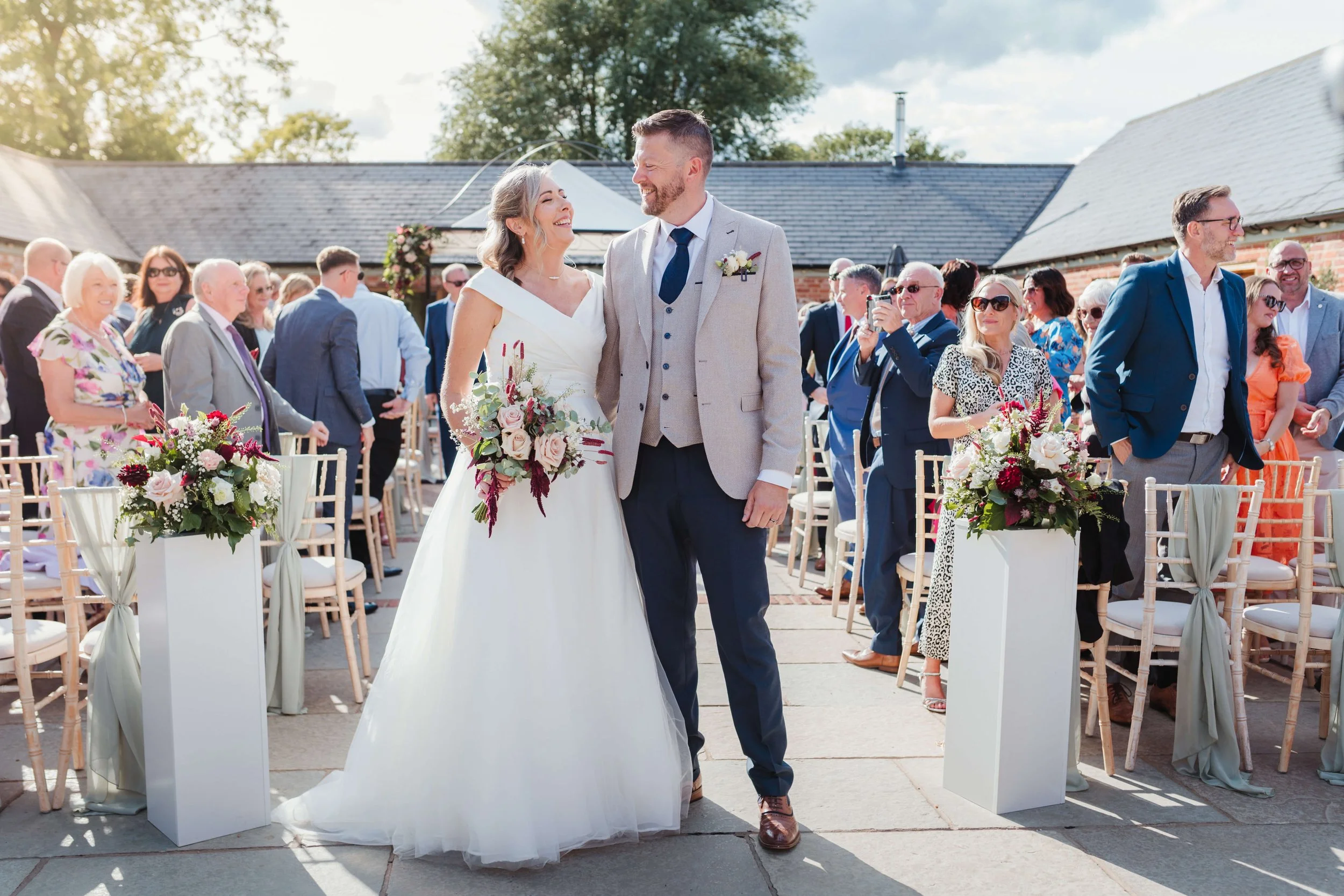 A bride and groom at their wedding ceremony, smiling at each other, surrounded by seated guests and floral decorations outdoors on a sunny day.