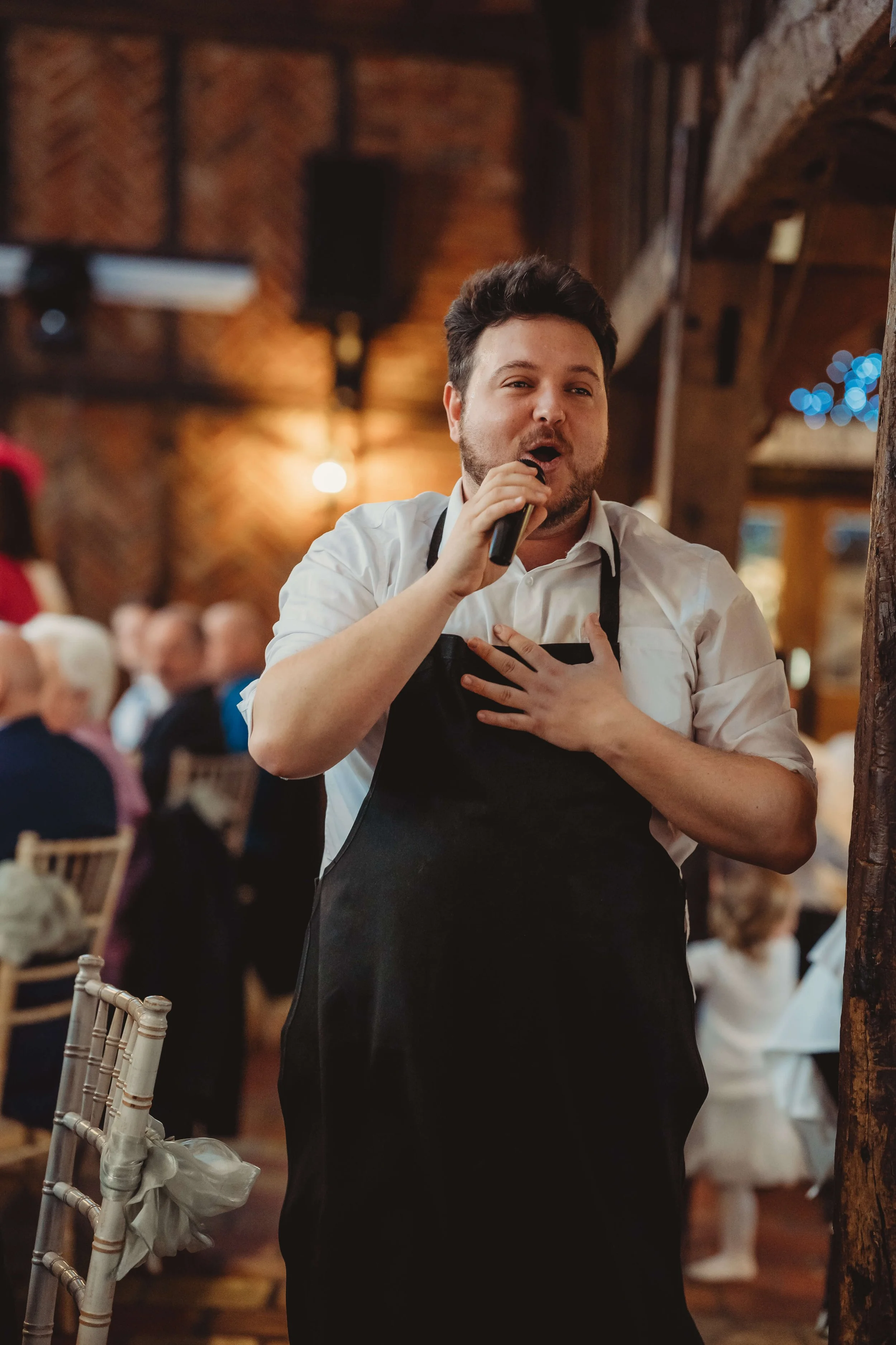 A man in a white shirt and black apron singing into a microphone with a hand on his chest, at a celebration or event in a rustic venue with wood beams and warm lighting.