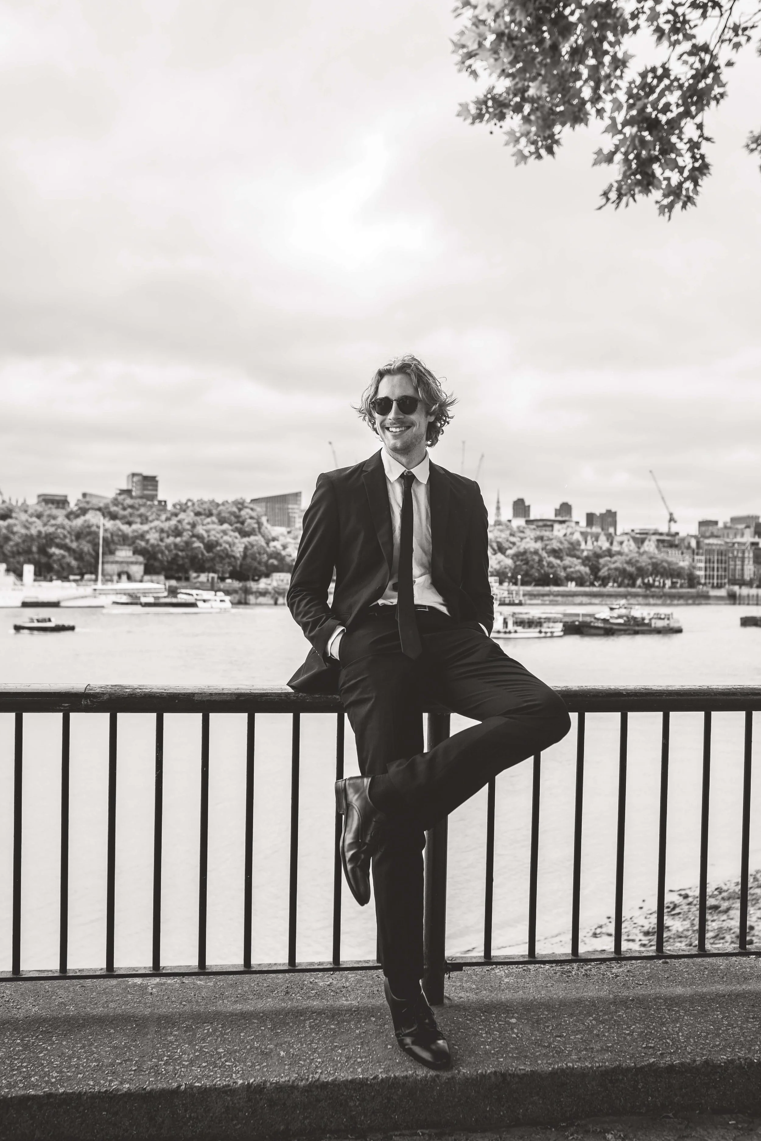 A man in a suit sitting on a railing near a river, smiling and wearing sunglasses, with a city skyline and boats in the background, in black and white.