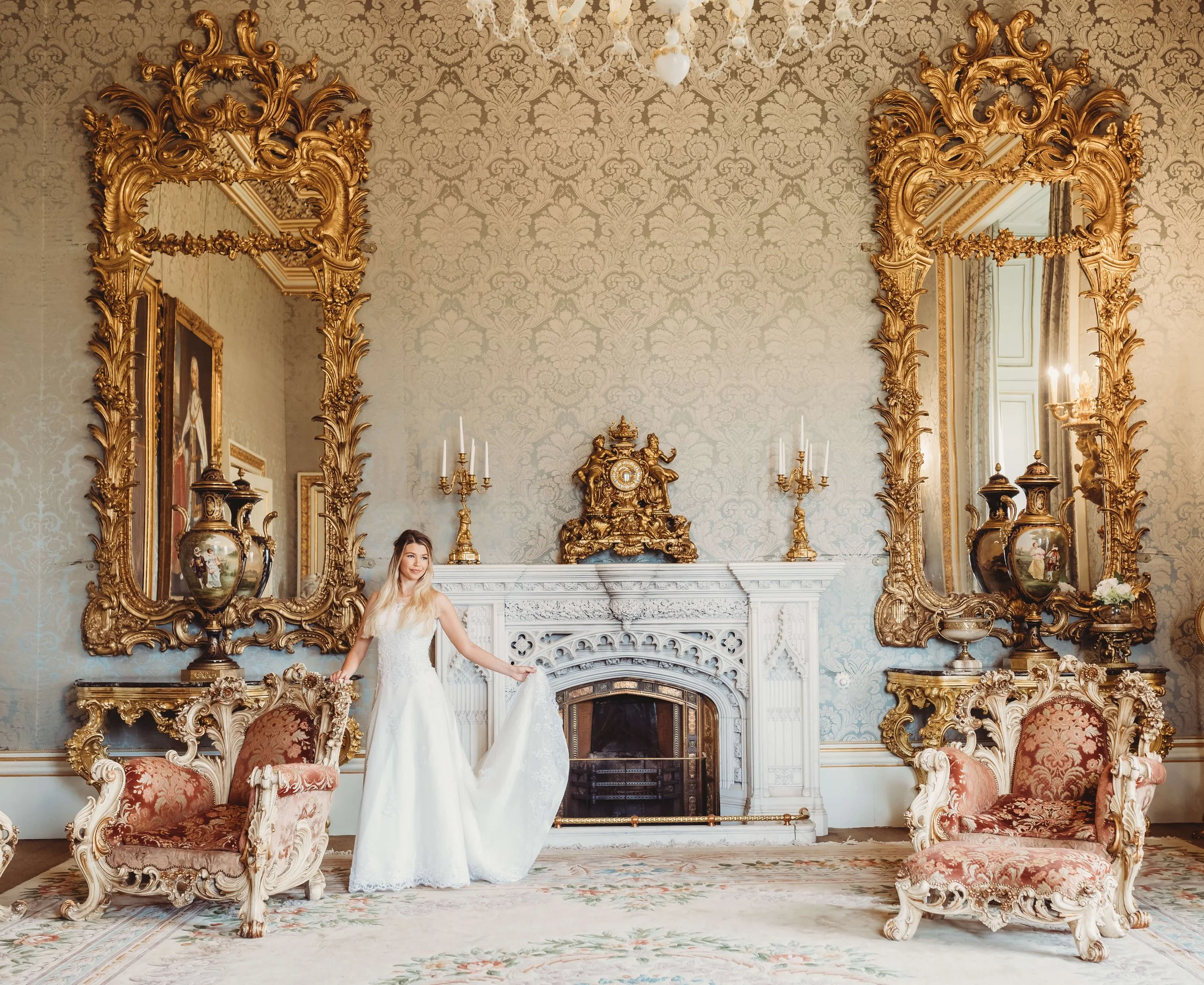 A woman in a white wedding dress standing in an opulent, historically decorated room with ornate gold-framed mirrors, a fireplace with an elaborate clock, and antique chairs. The room features intricate wall patterns and classic decor.