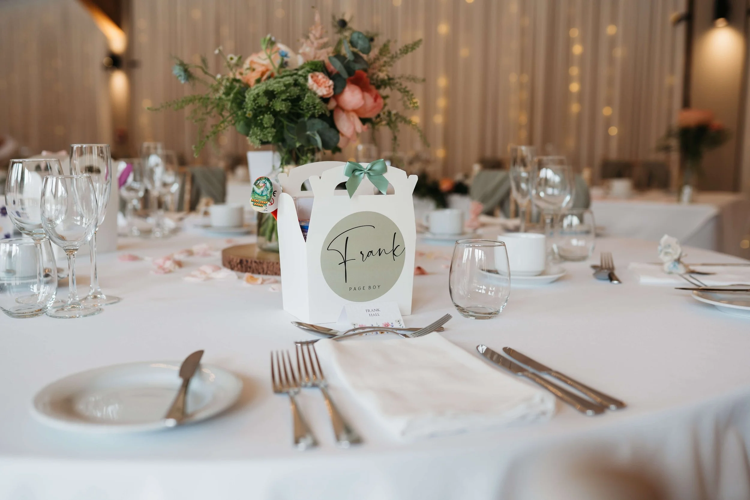 Elegant banquet table setup with a white tablecloth, glassware, silverware, a floral centerpiece, and a personalized name card for Frank. The background features warm lighting and draped curtains.