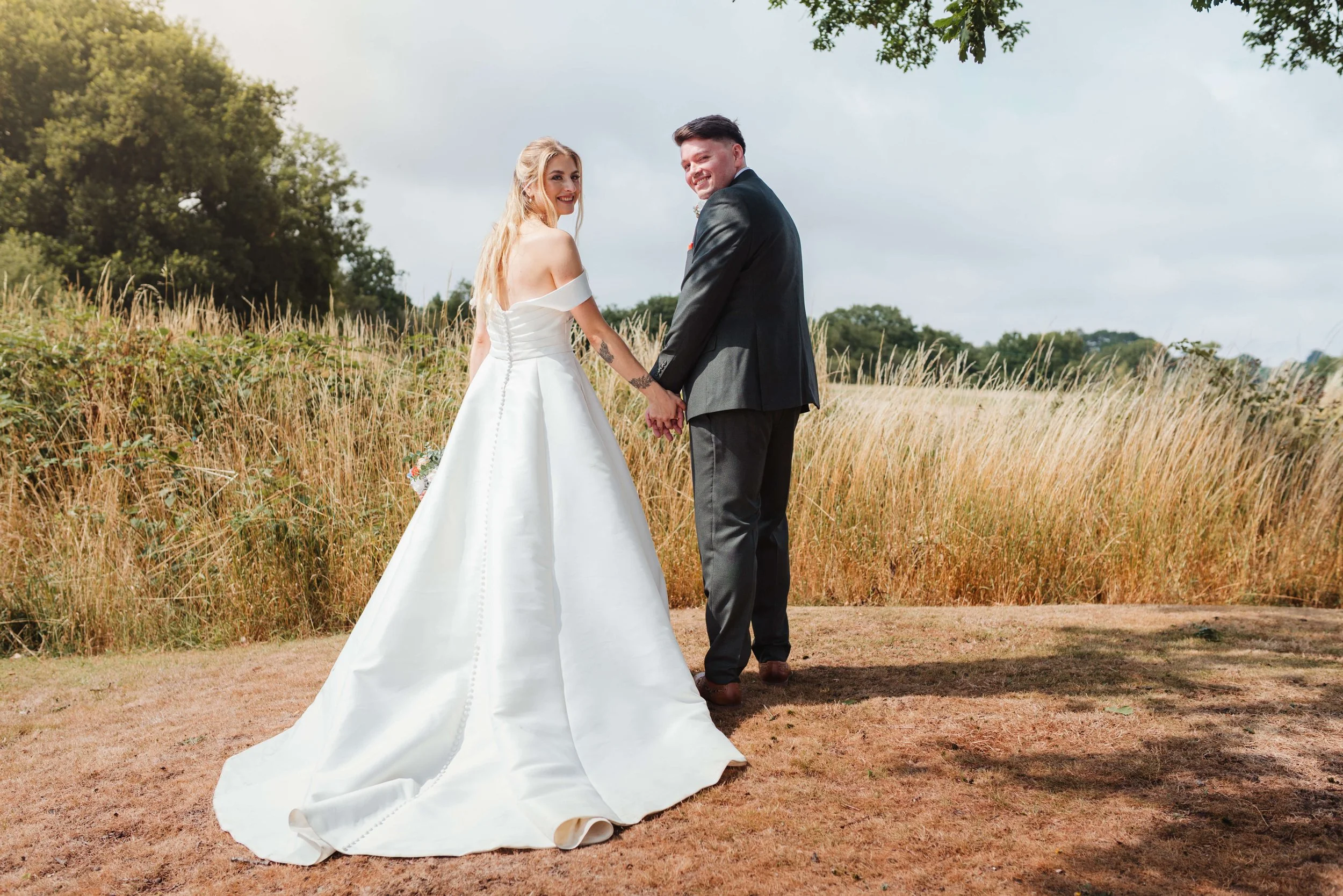 A bride and groom holding hands and smiling in a field with tall grass and trees in the background.