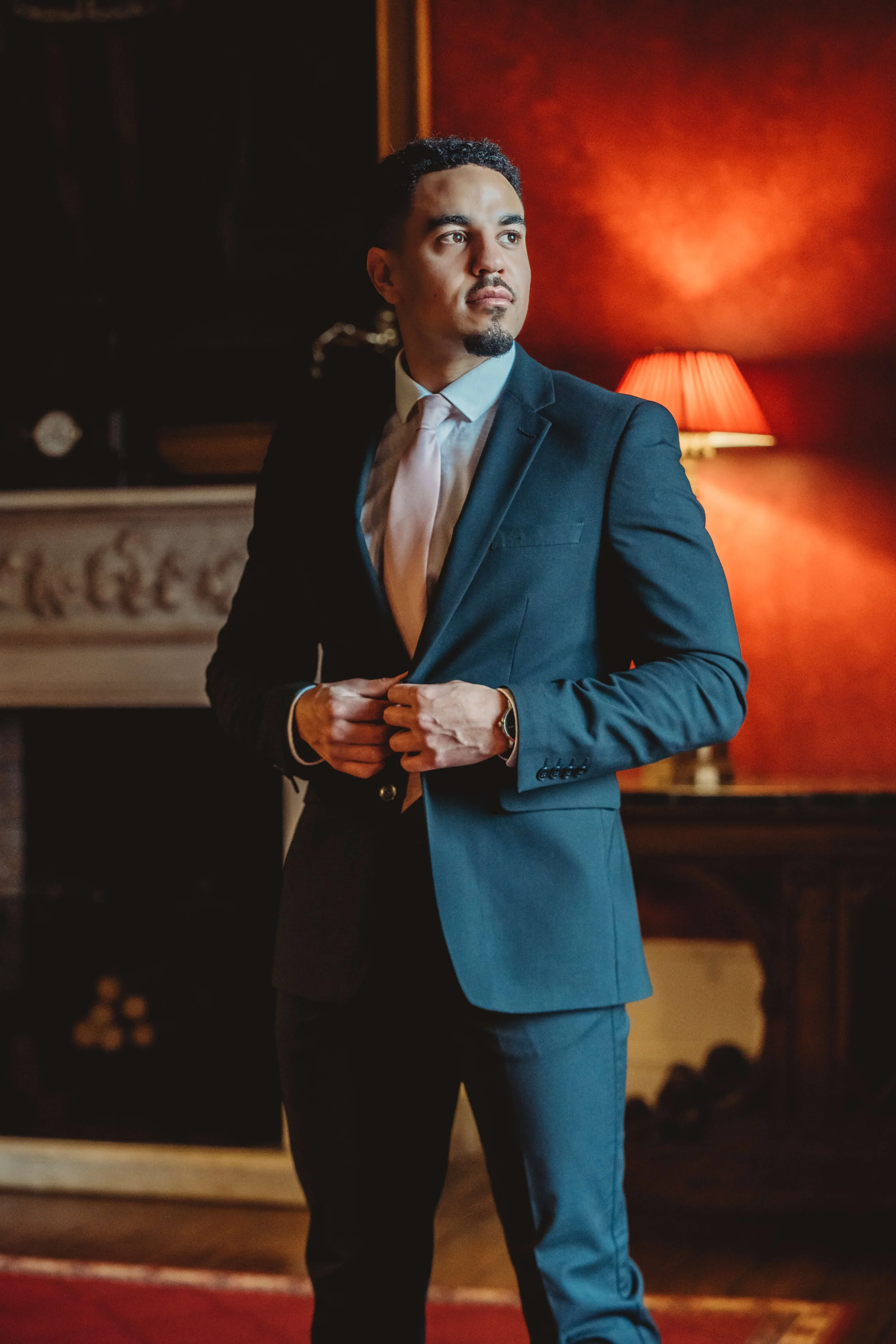 A man in a dark suit adjusting his jacket in a dimly lit room with a red wall and a lamp in the background.