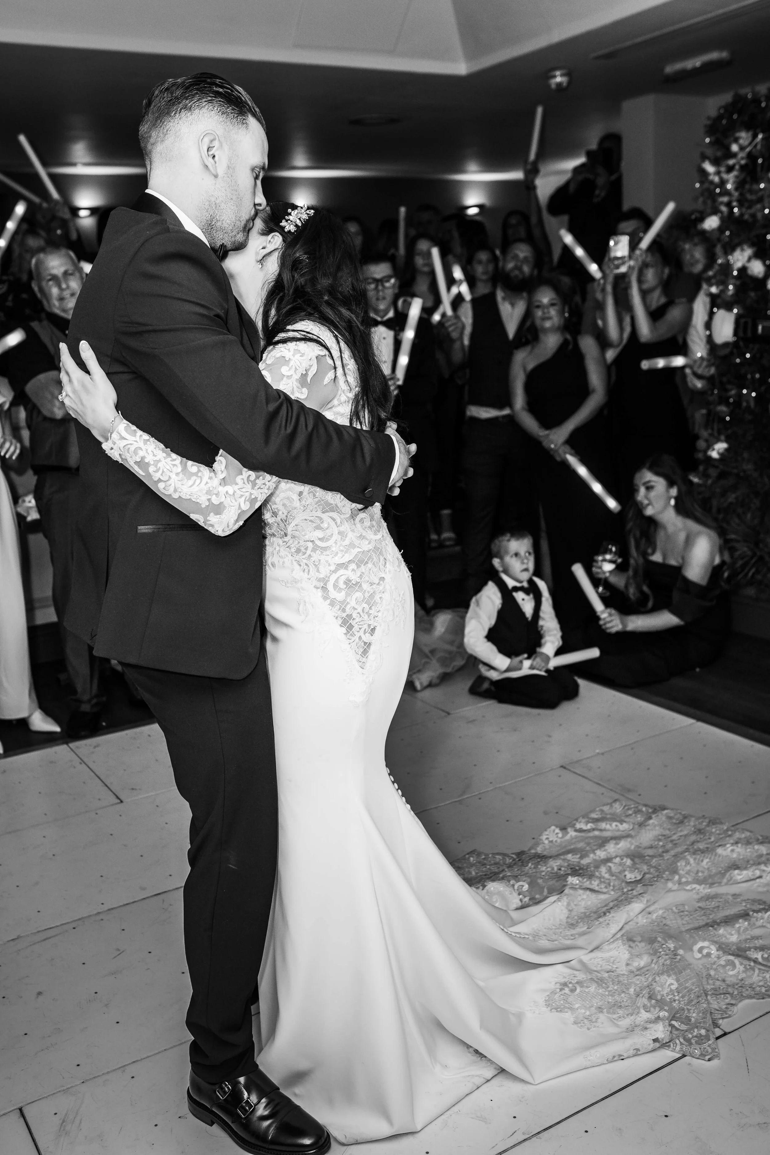 A black and white photo of a bride and groom sharing their first dance at their wedding reception, surrounded by guests watching and celebrating.