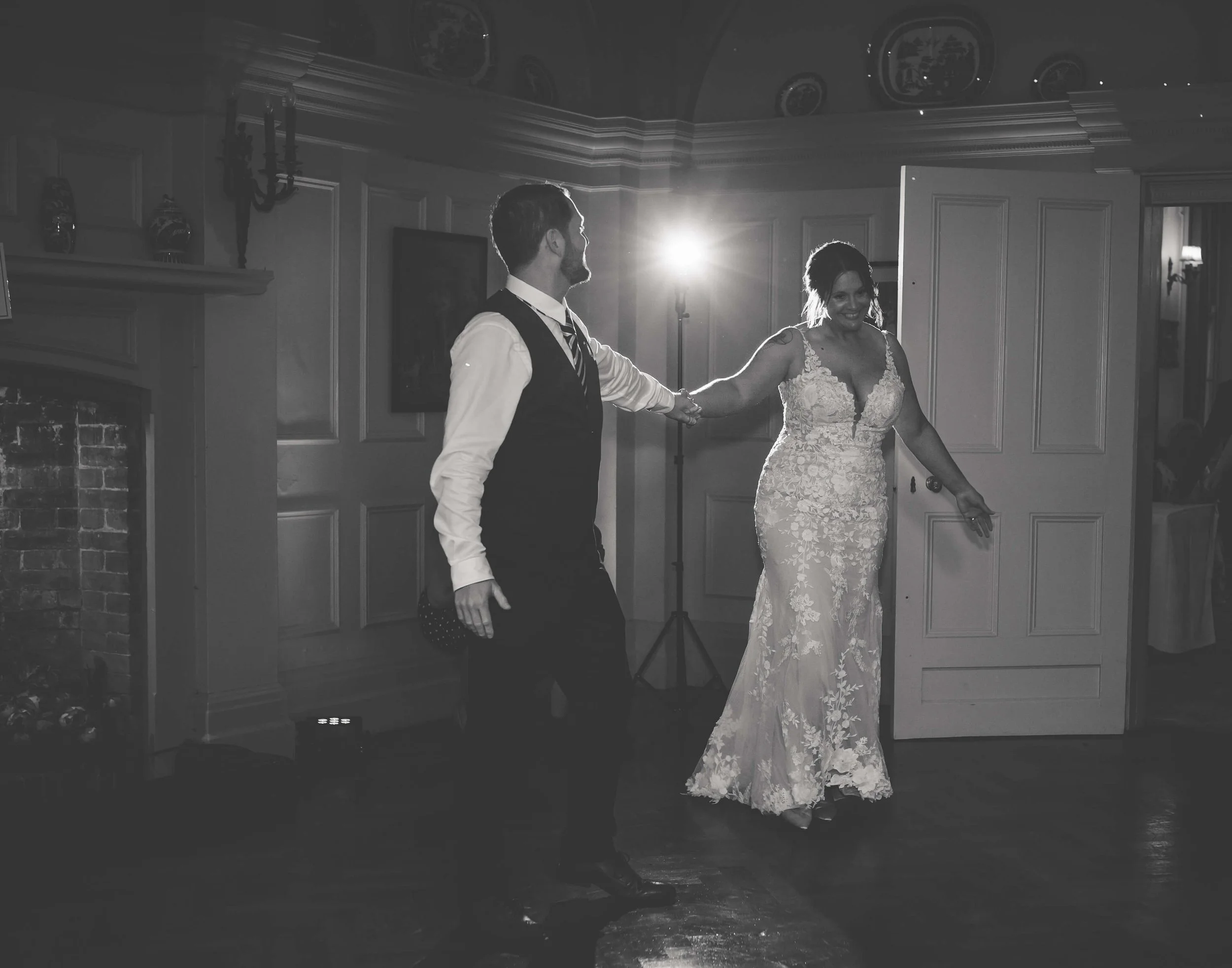 A bride and groom dancing at their wedding reception in an elegant room with a fireplace, decorative plates on the wall, and a bright light in the background.