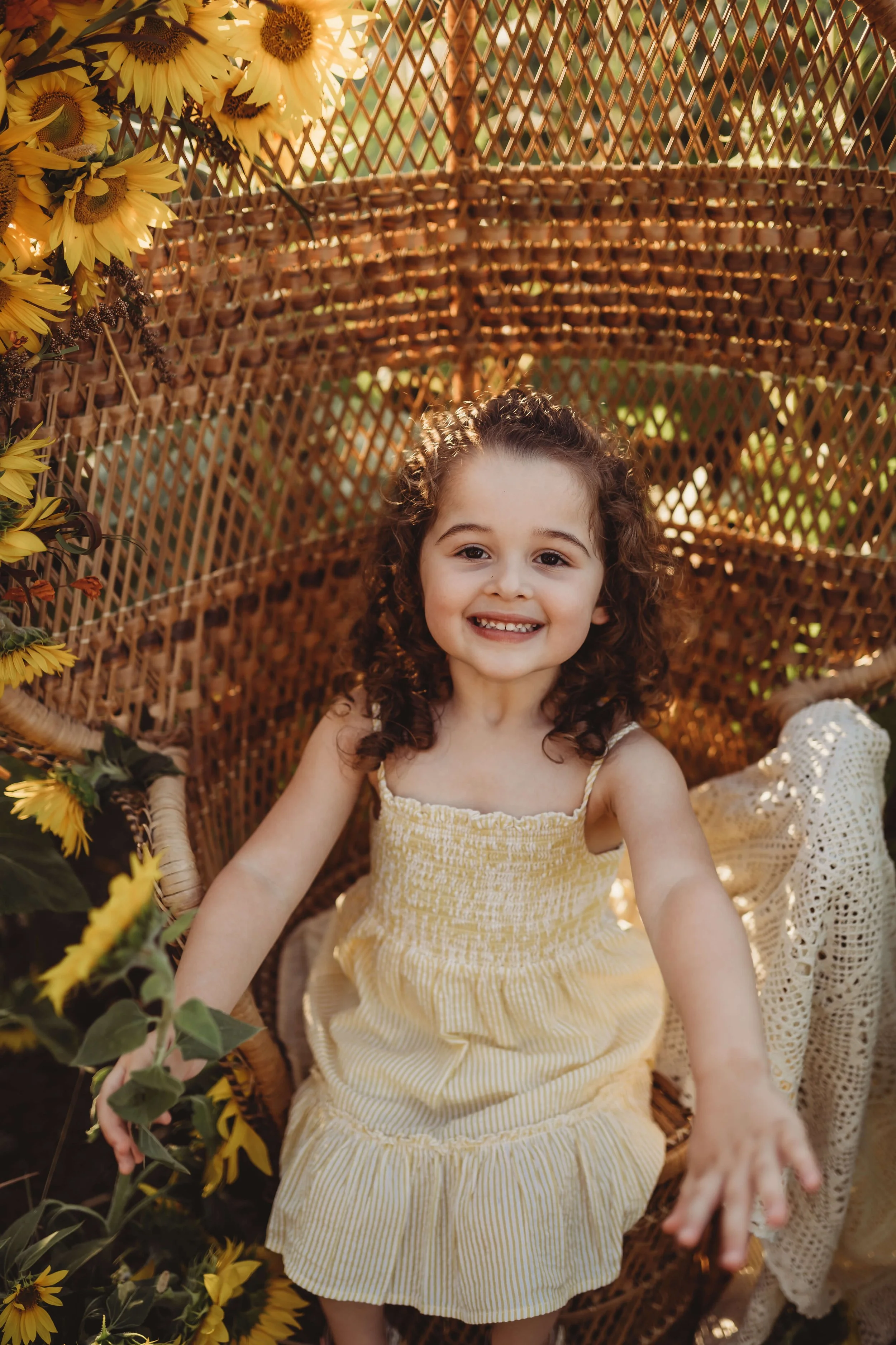 A young girl with curly hair smiling and reaching out, seated inside a wicker chair surrounded by sunflowers