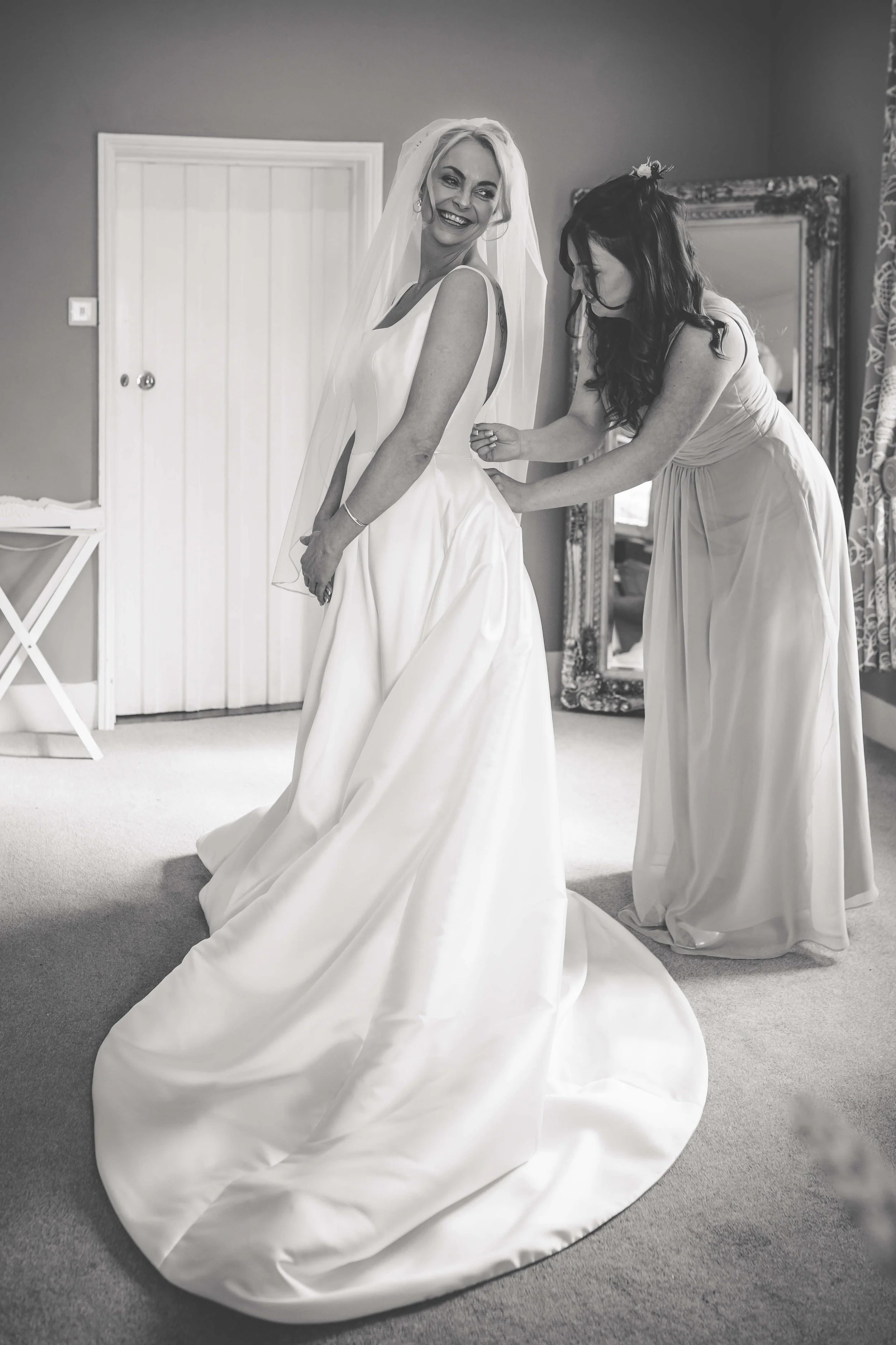 A bride in a wedding dress smiling as her bridesmaid assists her in getting ready in a room with a mirror and curtains.