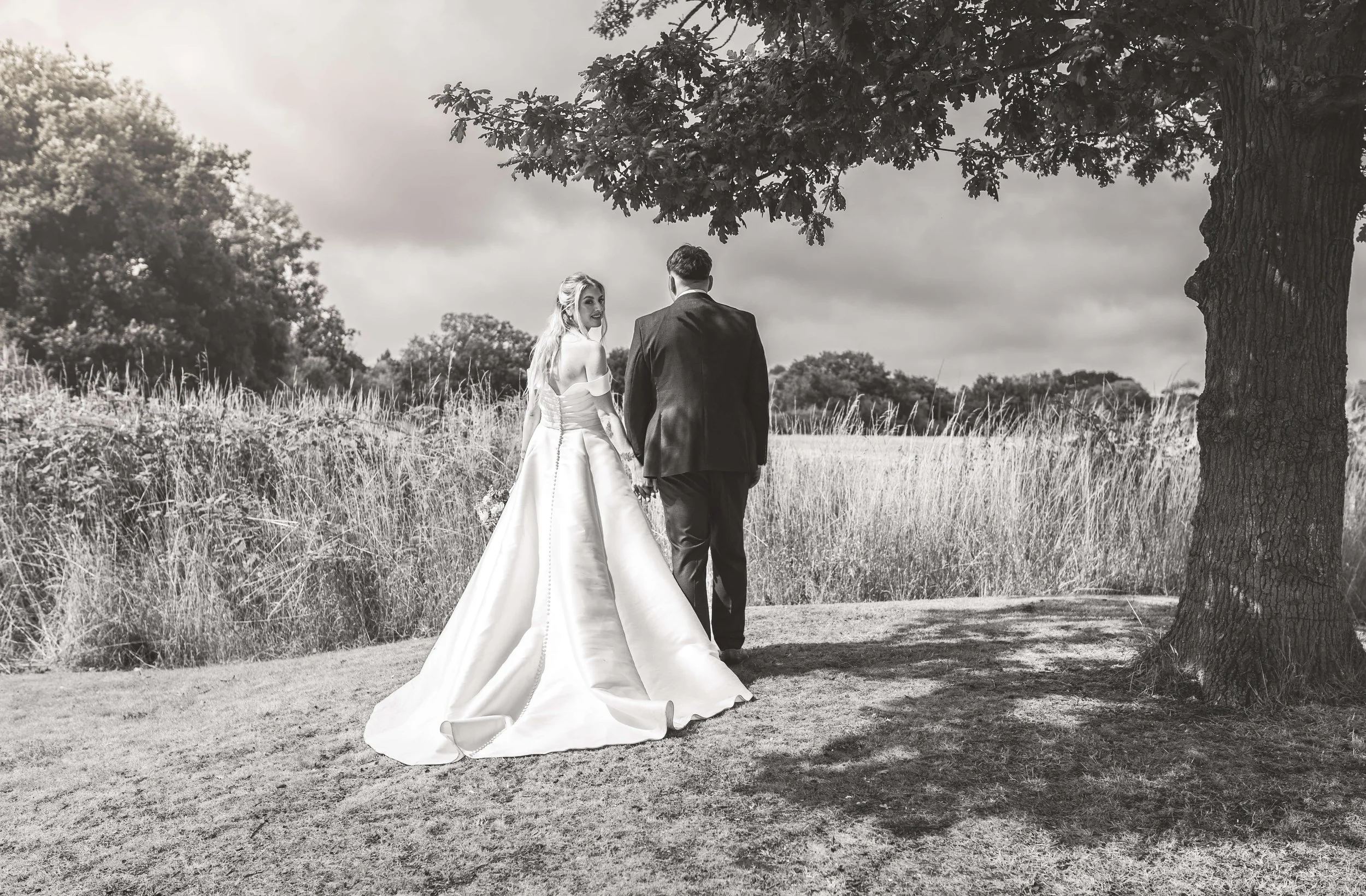 Black and white photograph of a bride and groom walking hand in hand outdoors, under a large tree, in a grassy field with a cloudy sky.