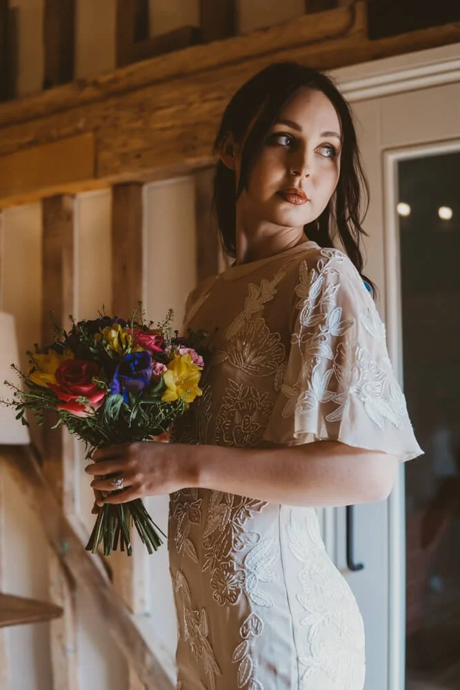 A woman in a white embroidered dress holding a colorful bouquet of flowers, standing indoors near a window at night.